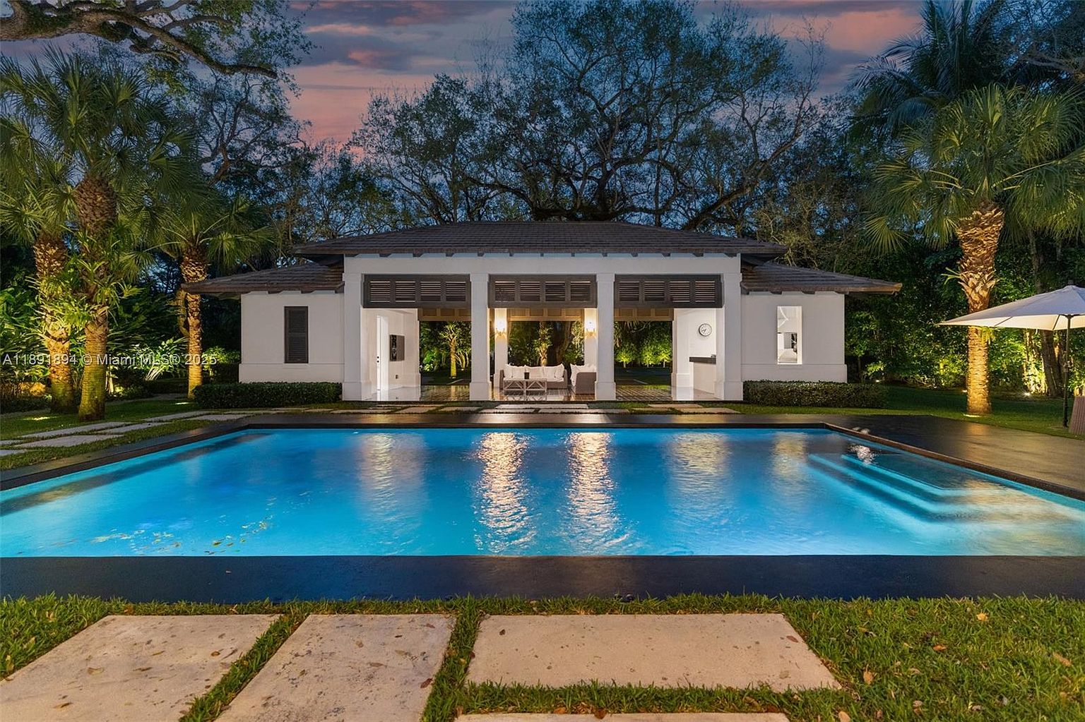 This image showcases a luxurious pool area at dusk, featuring a large rectangular pool with illuminated water. A modern pavilion with open sides and comfortable seating sits at the pool's edge, creating an inviting space for relaxation. The surrounding landscape includes mature trees and manicured lawns, enhancing the property's appeal.