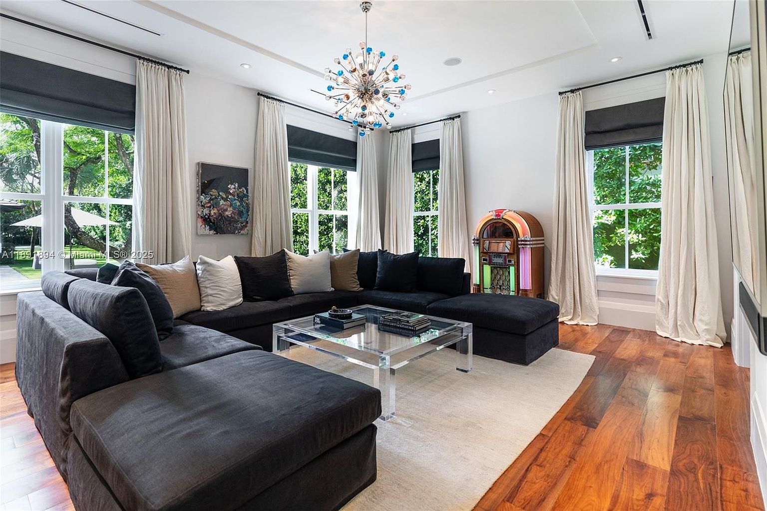 This is an interior shot of a living room featuring a large, dark gray sectional sofa with multiple pillows, a clear acrylic coffee table with books, and a light-colored area rug. The room has hardwood floors, white walls, and several windows with both dark gray shades and light-colored curtains. A colorful jukebox stands near one of the windows, and a modern chandelier hangs from the ceiling, adding a touch of elegance.