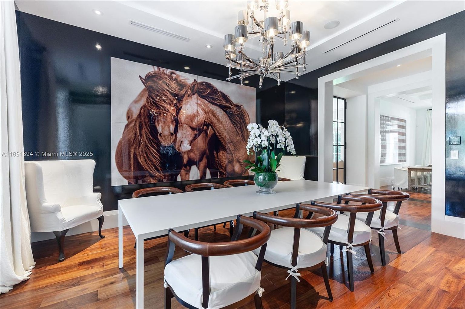 This is an interior shot of a dining room featuring a long white table with seating for eight. The chairs have dark wood frames and white cushions. A large piece of art depicting horses adorns the wall, and a modern chandelier hangs above the table. The room has dark walls and hardwood floors, creating a sophisticated and elegant atmosphere.