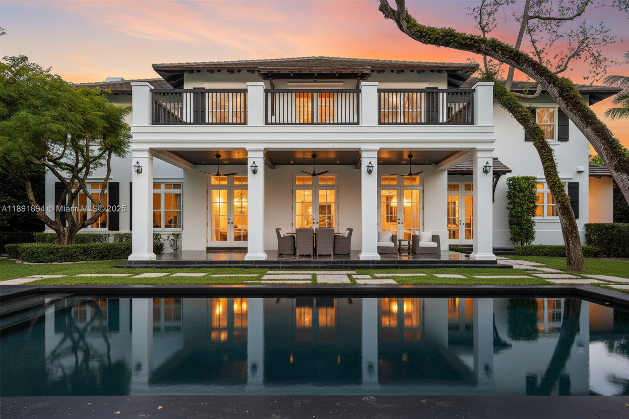 This image showcases the rear exterior of a luxurious home with a focus on its outdoor living space. A large swimming pool reflects the warm glow of the house lights, which are complemented by a spacious covered patio with elegant columns and an upper balcony. The well-manicured lawn and mature trees enhance the property's appeal.
