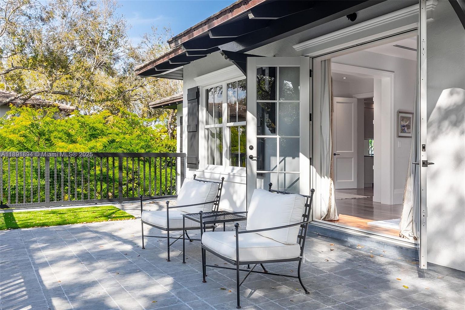 This image showcases a charming patio area, featuring two wrought iron chairs with white cushions and a small table, creating an inviting outdoor seating space. The patio is paved with gray stone tiles and overlooks a lush green lawn and garden area, framed by a dark railing. French doors lead into the home, suggesting a seamless indoor-outdoor living experience.
