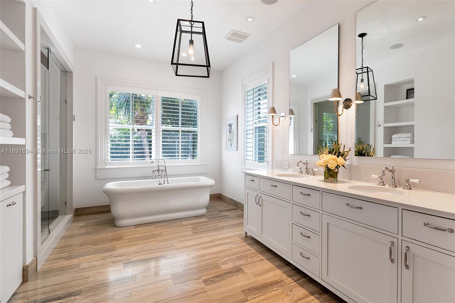 This is a bright and spacious primary bathroom featuring a freestanding bathtub positioned under a window with plantation shutters. A double vanity with white cabinetry and light countertops is complemented by large mirrors and stylish sconces. The flooring appears to be wood-look tile, adding warmth to the room.