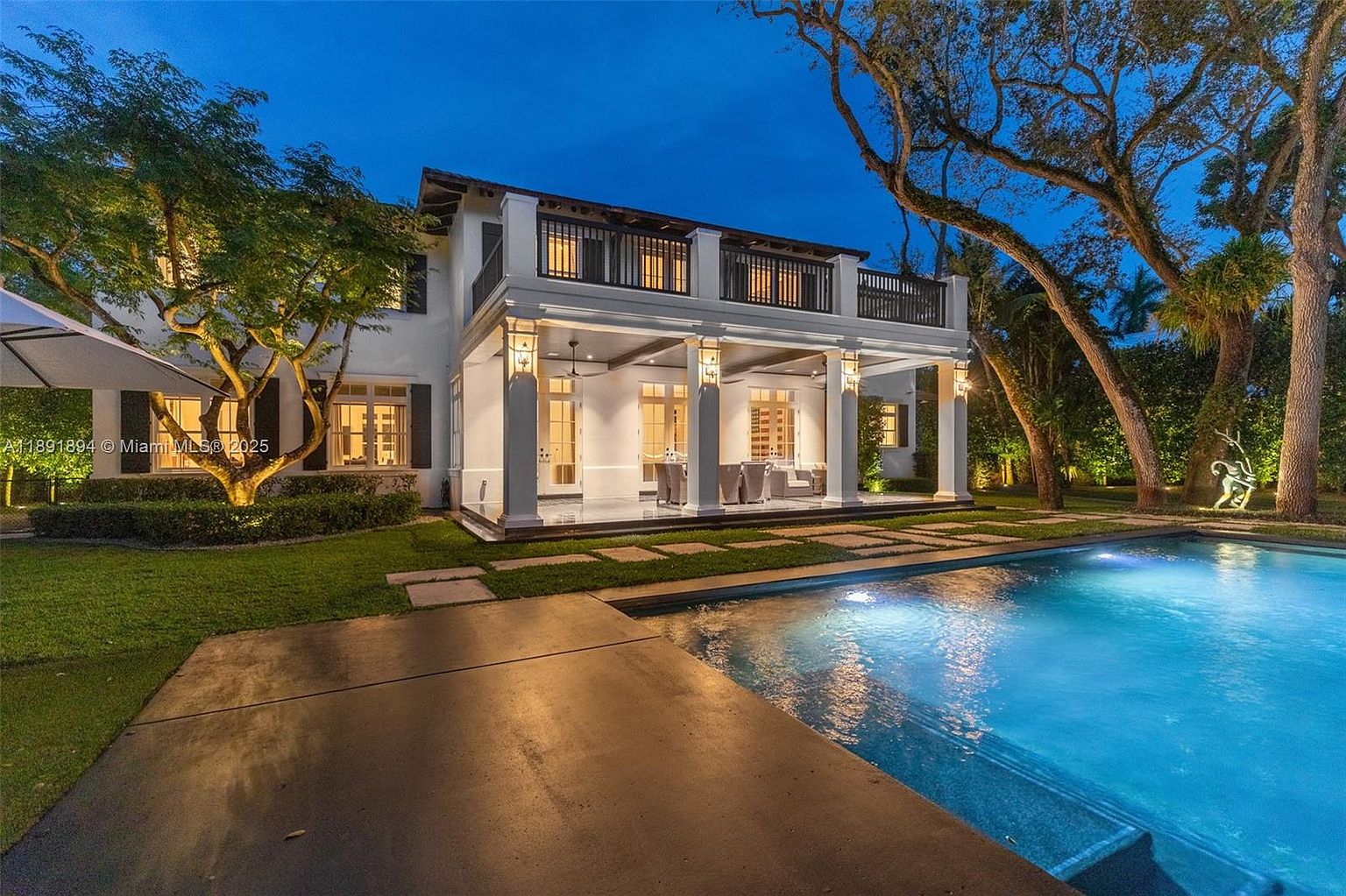 This image showcases the rear exterior of a luxurious two-story home, highlighting a stunning pool area. The architecture features white columns supporting a covered patio and a balcony above. The pool is illuminated, creating a serene and inviting atmosphere, while mature trees and manicured landscaping add to the property's appeal.