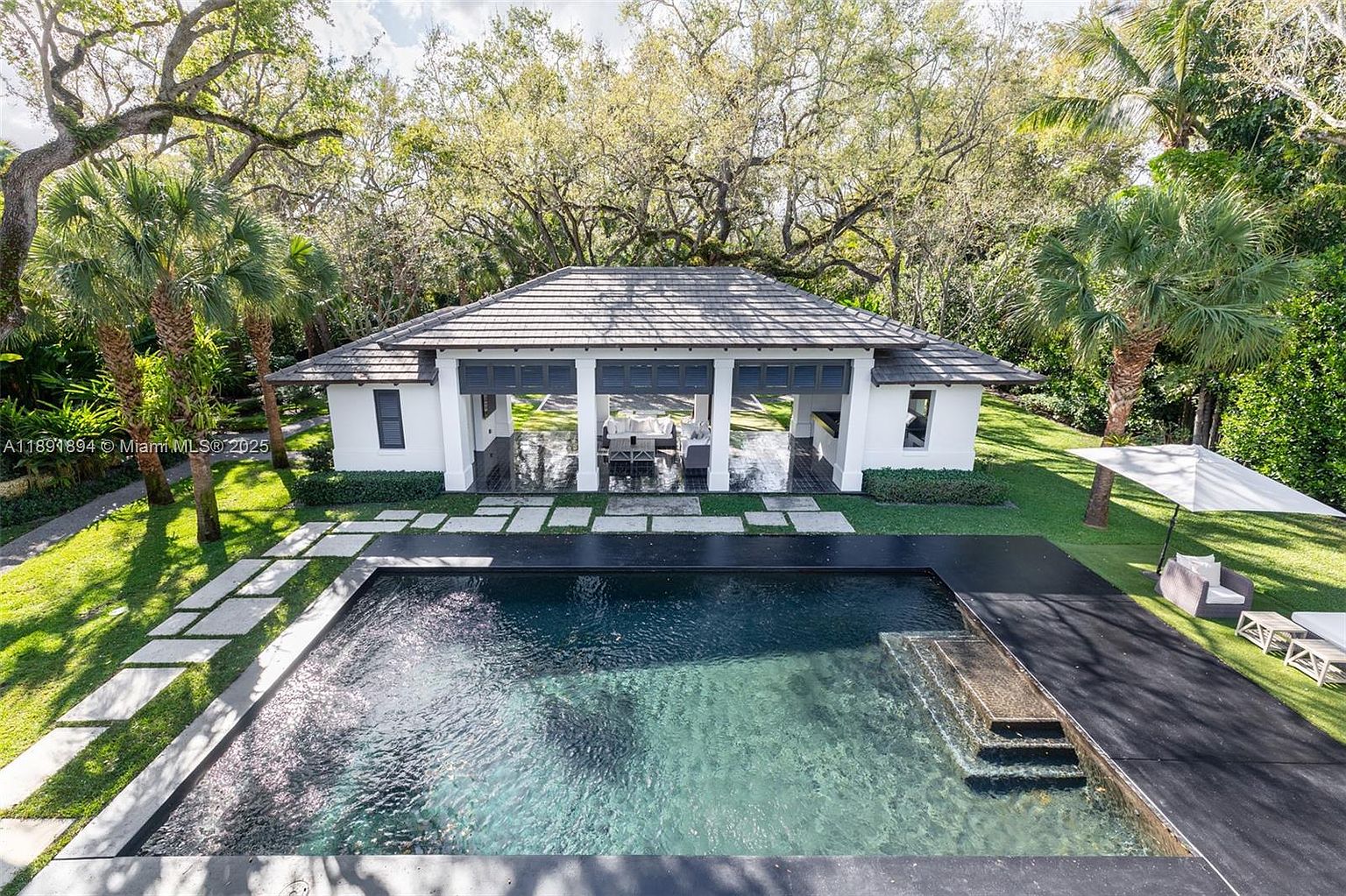 This image showcases a luxurious backyard pool area. The pool features a dark, reflective surface and is surrounded by a modern deck and stepping stones leading to a covered outdoor living space. Lush greenery and mature trees provide privacy and a serene backdrop, enhancing the property's appeal.