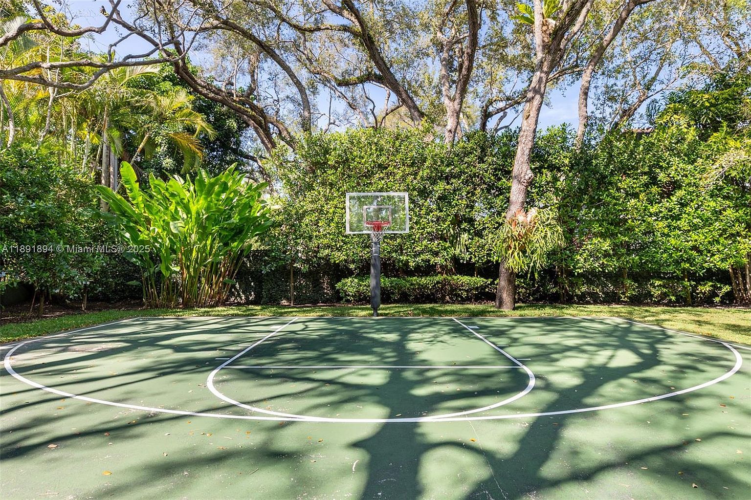 The image showcases a well-maintained outdoor basketball court surrounded by lush greenery, including trees and hedges, creating a private and serene atmosphere. The court features clear white lines on a green surface, with a basketball hoop centered at the far end. The scene evokes a sense of recreation and relaxation within a beautifully landscaped setting.