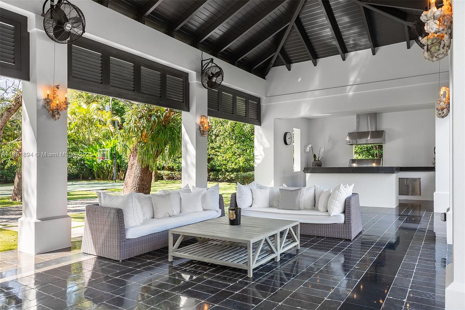 This image showcases a luxurious outdoor living space featuring two wicker sofas with white cushions arranged around a light gray wooden coffee table. The patio has a black tiled floor and is partially covered by a dark wood ceiling, creating a shaded and elegant area for relaxation and entertainment. An outdoor kitchen is visible in the background, enhancing the functionality of the space.