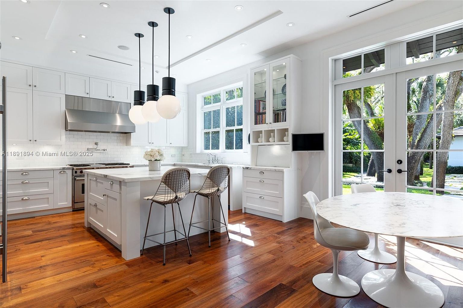 This is a bright and airy kitchen featuring white cabinetry, a stainless steel range hood, and a large island with a marble countertop. Modern pendant lights hang above the island, and a breakfast nook with a marble table and white chairs sits near a set of French doors leading to the outside. The hardwood floors add warmth to the space, creating an inviting atmosphere.