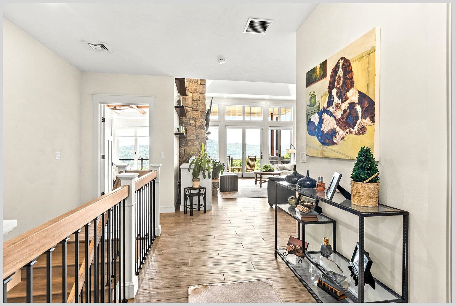 This perspective captures a bright, open-concept hallway leading toward a sun-drenched living area with expansive windows. To the right, a sleek metal console table displays decorative items beneath a large, vibrant painting of dogs, while a staircase with a dark metal railing borders the left side. The space features light-toned wood-look flooring and a stone-clad chimney breast that anchors the transition between the hallway and the main living space.