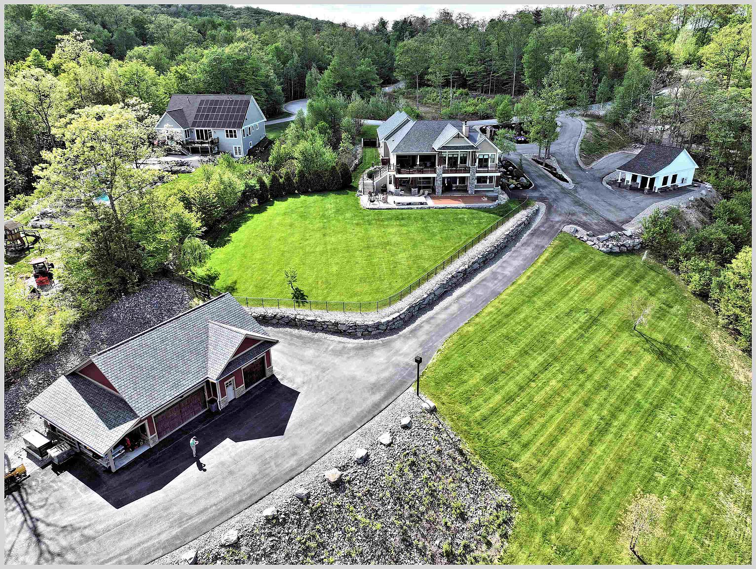 This high-angle aerial view captures a sprawling residential property nestled within a lush, wooded landscape. The estate features a primary multi-level residence with a spacious deck, a separate detached garage, and a smaller outbuilding, all connected by paved driveways. Expansive, well-manicured green lawns and stone retaining walls define the outdoor living spaces, creating a serene and private atmosphere.