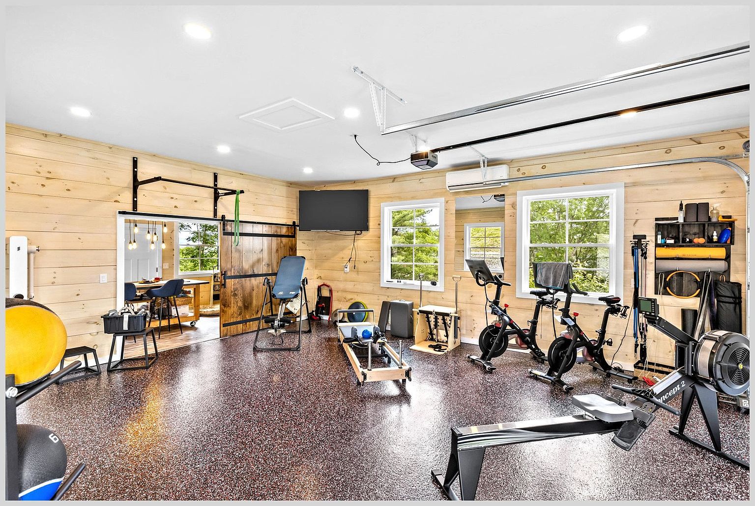This spacious home gym features light wood-paneled walls and durable, speckled flooring, creating a bright and functional workout environment. The room is well-equipped with a variety of fitness gear, including a rowing machine, two stationary bikes, a Pilates reformer, and an inversion table. A sliding barn door provides a rustic touch while offering a glimpse into an adjacent dining area, and recessed lighting keeps the space evenly illuminated.