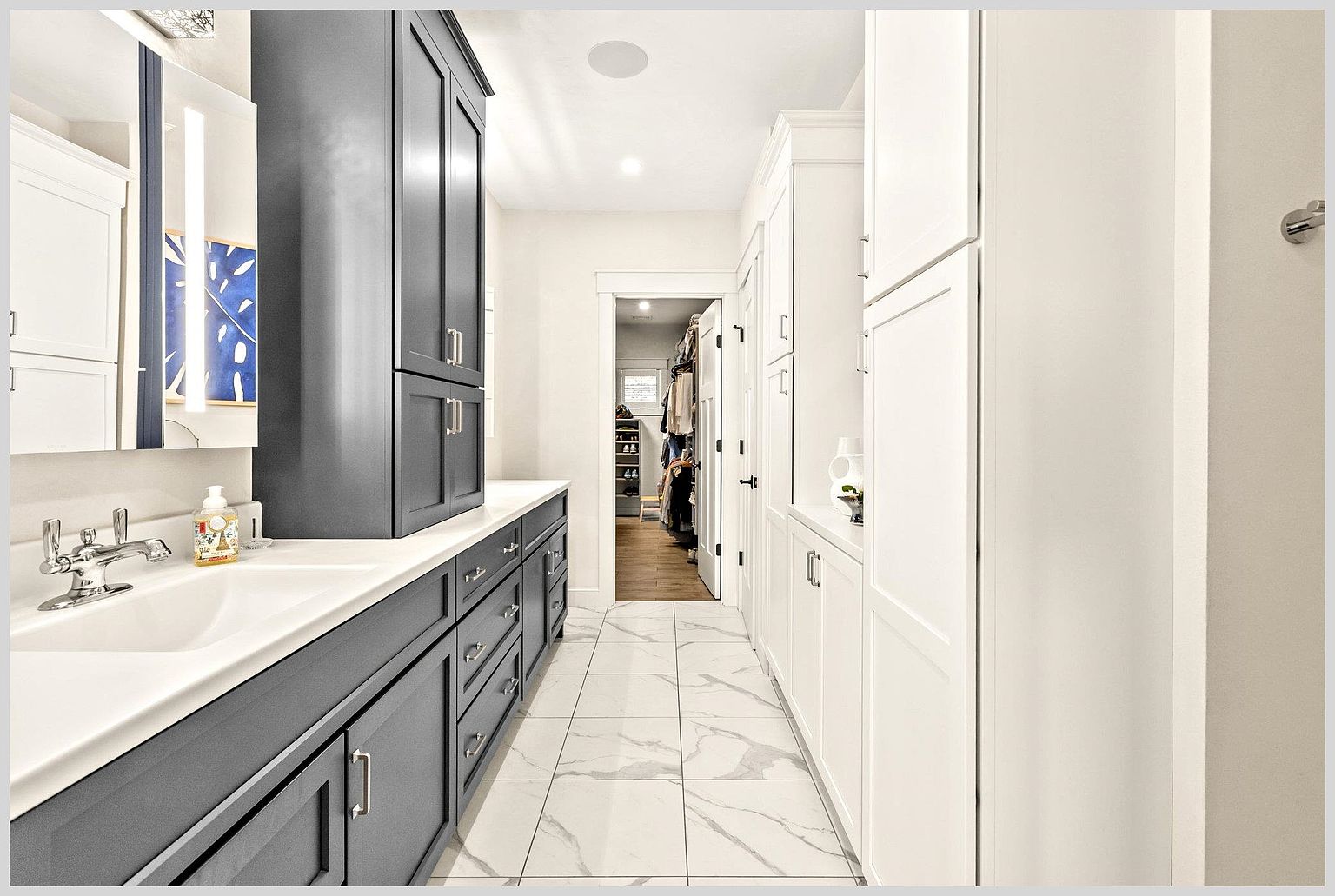 This primary bathroom features a striking contrast between dark charcoal cabinetry on the left and crisp white cabinetry on the right. The space is anchored by light-colored, large-format patterned tile flooring and leads directly into a walk-in closet visible through the doorway. The clean lines and modern fixtures create a sophisticated, high-end aesthetic suitable for a luxury home.