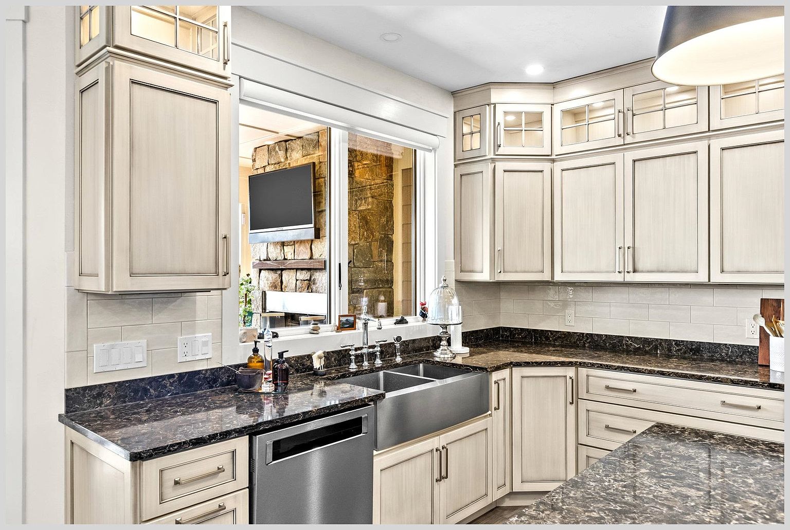 This kitchen features light-toned cabinetry with a subtle distressed finish, paired with dark, patterned stone countertops and a stainless steel farmhouse sink. A large window above the sink provides a view into an adjacent room with a stone-clad chimney breast and a mounted television. The space is well-lit with recessed lighting and a pendant fixture, creating a warm and inviting atmosphere.