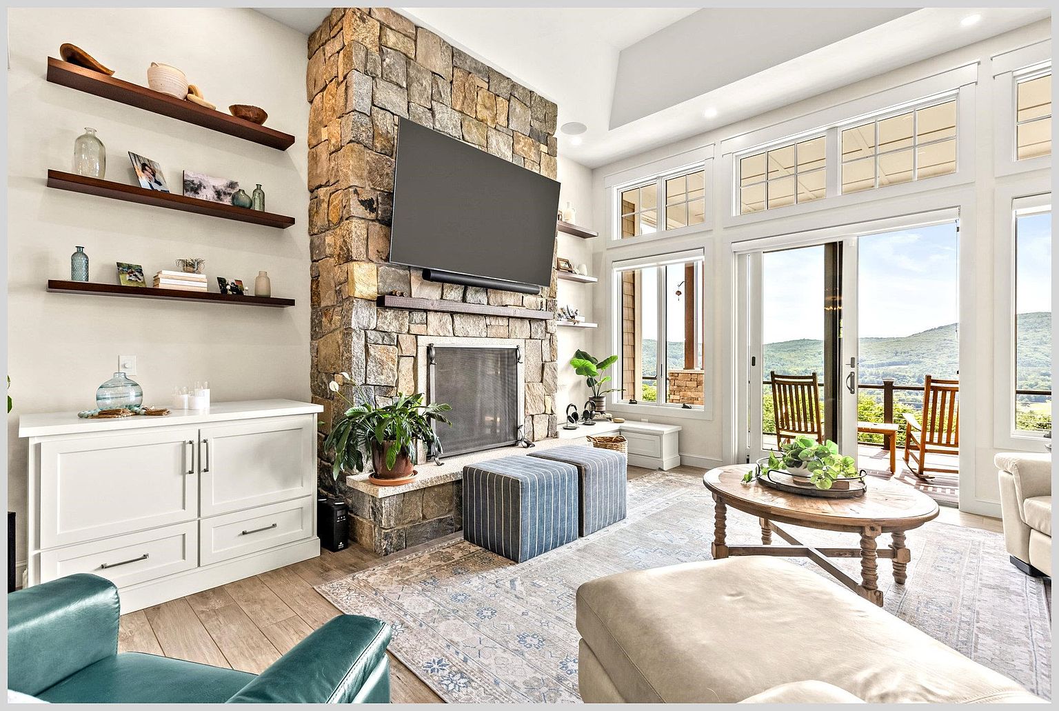 This inviting living room features a prominent stone-clad chimney breast that serves as the focal point, complemented by built-in shelving and a white storage console. Large windows and glass doors provide abundant natural light and expansive views of the surrounding landscape, while a patterned area rug anchors the seating area. The space is furnished with a mix of comfortable seating, including a plush ottoman and a pair of blue-striped stools, creating a warm and relaxed atmosphere.