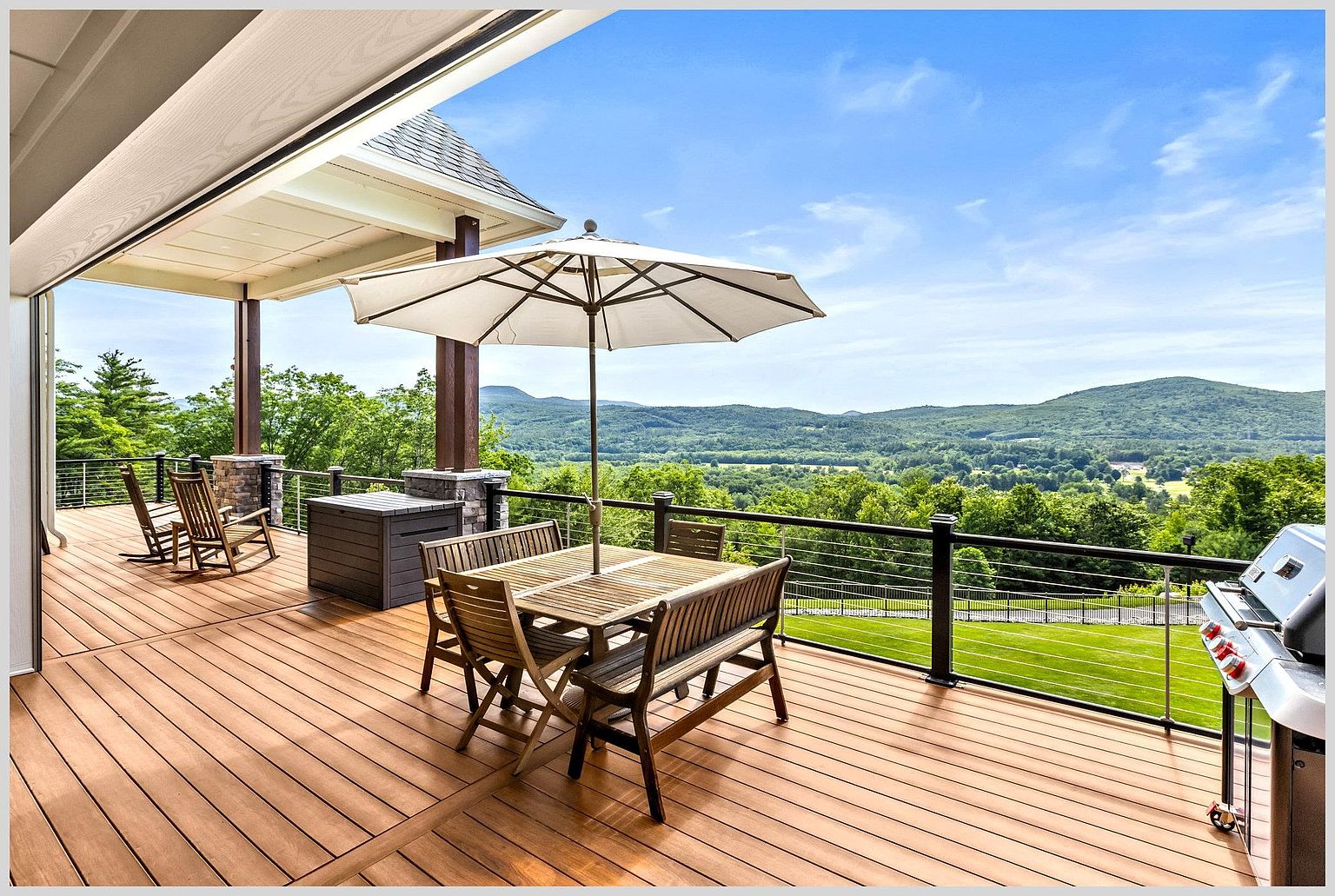 This expansive, elevated wooden deck offers a panoramic view of rolling green hills and a lush valley under a bright blue sky. The space is furnished with a dining set shaded by a large umbrella and a pair of rocking chairs, all positioned to take advantage of the scenic vista. A stainless steel grill sits to the right, while a sleek cable railing ensures an unobstructed connection to the natural landscape beyond.