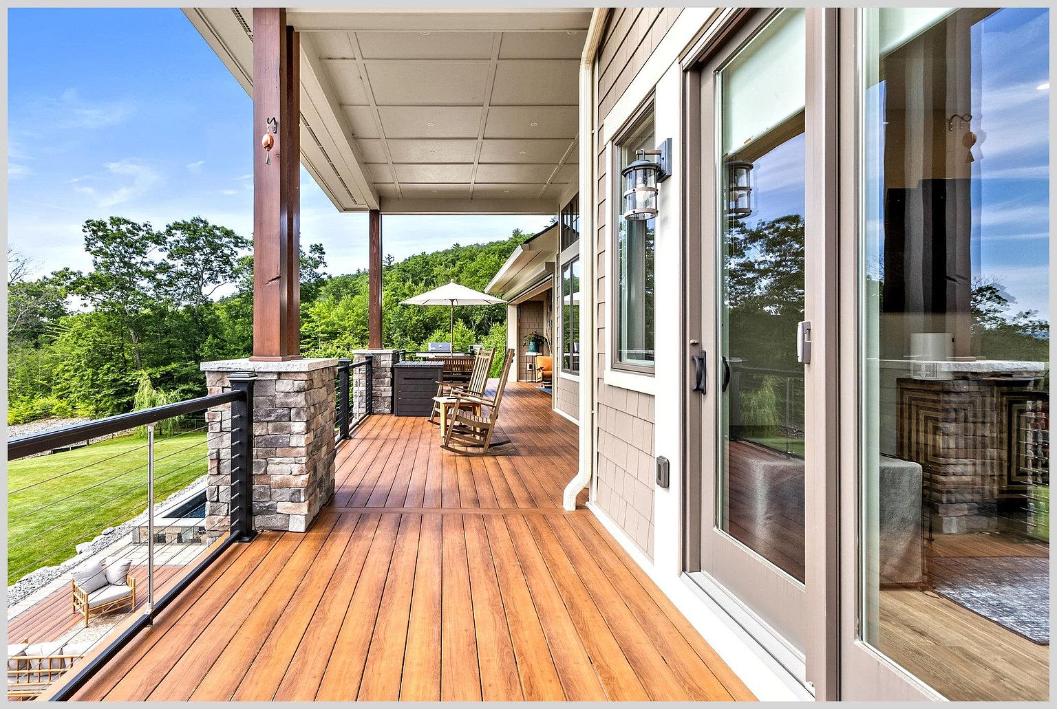This spacious covered deck features warm-toned wood decking and a stone-based railing system with thin metal cables, offering an unobstructed view of the surrounding lush green landscape. The area is furnished with wooden rocking chairs and a patio umbrella, creating a comfortable outdoor living space that seamlessly connects to the interior through large glass sliding doors. The perspective captures the length of the deck, highlighting the transition between the sheltered seating area and the open, scenic backyard.