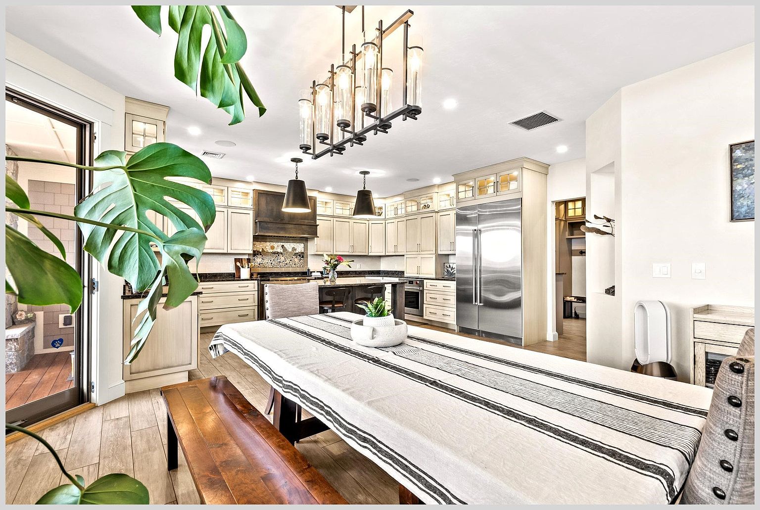 This spacious, open-concept kitchen features light-toned cabinetry, a large central island with bar seating, and high-end stainless steel appliances. A long dining table with a striped runner and a wooden bench sits in the foreground, while a large, modern chandelier hangs above the kitchen area. The room is bright and inviting, with a view through a glass door to an outdoor space and a large plant in the foreground adding a touch of greenery.