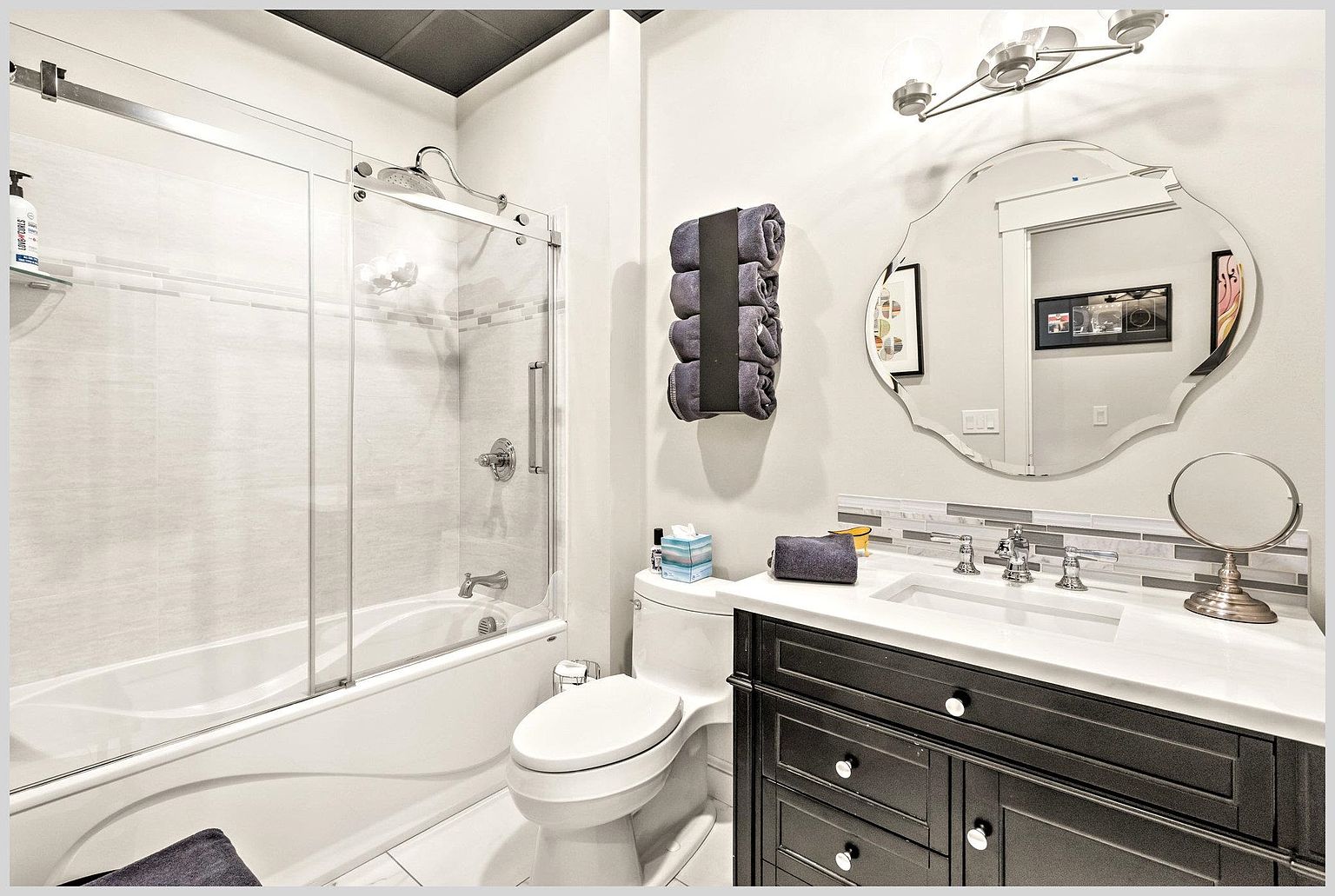 This well-appointed bathroom features a dark vanity with a white countertop, a scalloped-edge mirror, and a glass-enclosed shower-tub combination. The space is accented with a tiled backsplash, rolled towels, and modern lighting fixtures, creating a clean and polished aesthetic. The perspective is a straight-on shot capturing the vanity, toilet, and shower area in a single, cohesive view.