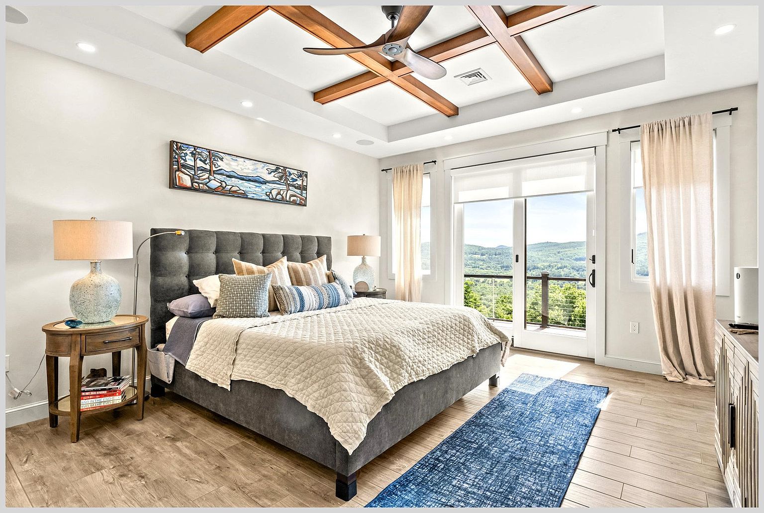 This primary bedroom features a sophisticated design with a tufted gray upholstered bed as the focal point, complemented by a coffered ceiling with exposed wood beams and a ceiling fan. A large glass sliding door provides access to a balcony with scenic mountain views, while light wood-toned flooring and a blue patterned area rug add warmth and texture to the space. The room is well-lit by recessed lighting and bedside lamps, creating a serene and inviting atmosphere.
