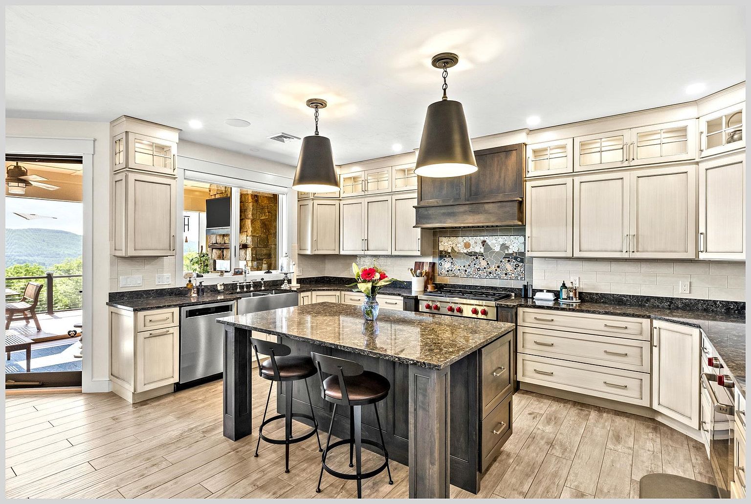 This spacious kitchen features a large central island with a dark base and stone countertop, complemented by two bar stools. The perimeter includes light-toned cabinetry, a stainless steel range with a custom hood, and a decorative backsplash featuring a rooster motif. Large windows provide a view of an outdoor deck and mountain scenery, while pendant lights hang above the island to create a warm, inviting atmosphere.