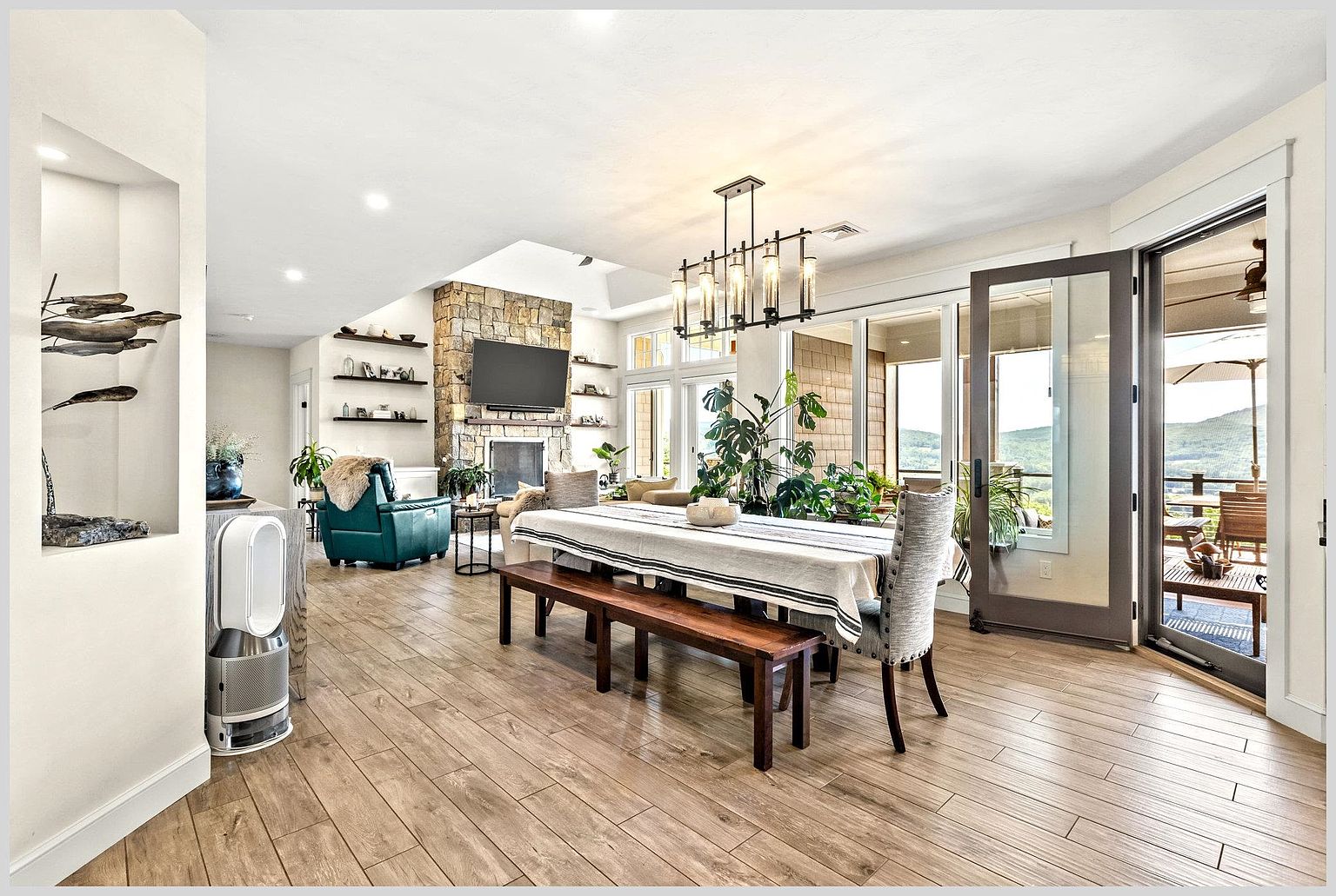 This open-concept dining area features a long wooden table with a bench and upholstered chairs, illuminated by a modern linear chandelier. The space flows seamlessly into a living area anchored by a stone-clad chimney breast and a television, with light wood-look flooring throughout. Large glass doors open to an outdoor deck, providing a bright, airy atmosphere with views of the surrounding landscape.