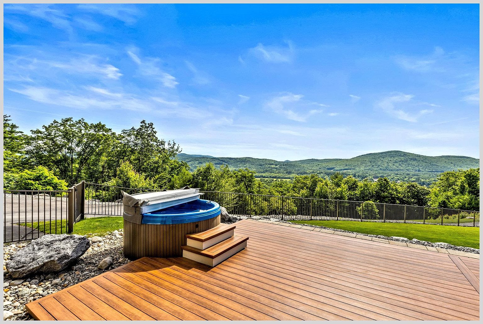 This expansive wooden deck features a built-in hot tub with a cover, providing a relaxing focal point against a backdrop of lush green trees and rolling mountains. A metal railing borders the deck, which overlooks a manicured lawn and the scenic landscape beyond. The bright, sunny atmosphere and elevated perspective create an inviting outdoor living space perfect for enjoying panoramic views.