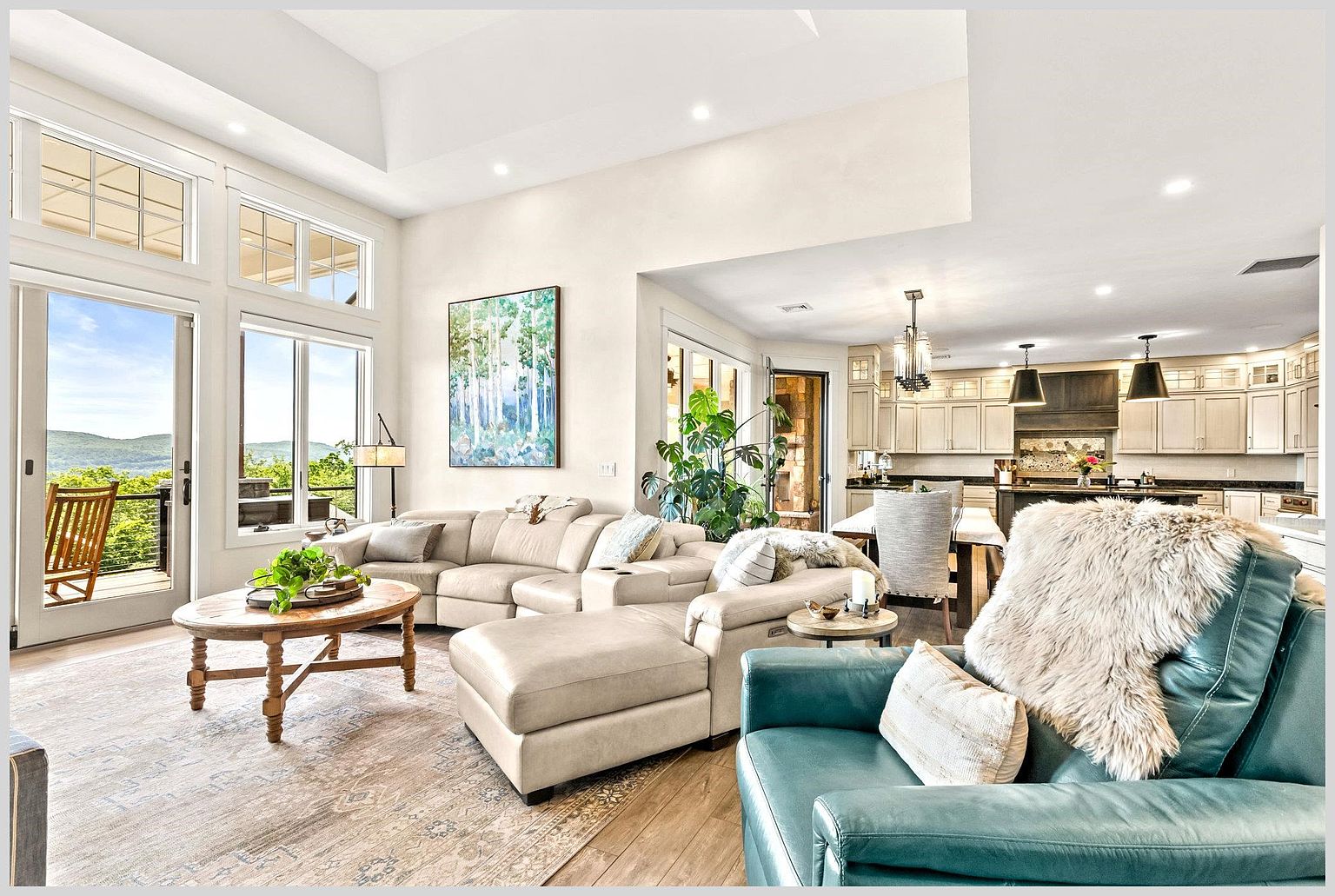 This open-concept living space features a neutral-toned sectional sofa arranged around a round wooden coffee table, with a vibrant teal armchair in the foreground adding a pop of color. Large windows and a glass door provide abundant natural light and a scenic view of the distant mountains. The room flows seamlessly into a spacious kitchen area, creating an inviting and airy atmosphere suitable for entertaining.