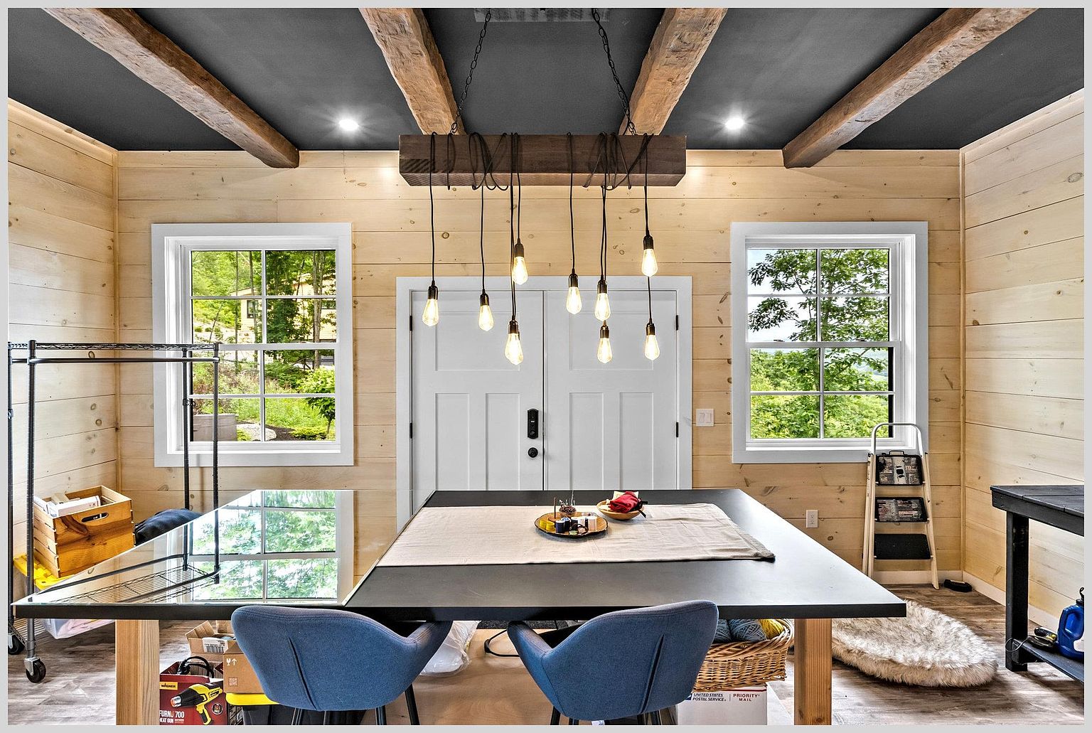 This rustic-inspired workspace features light wood-paneled walls and a dark ceiling accented by exposed wooden beams. A large central table serves as a primary workstation, illuminated by a unique multi-pendant light fixture suspended from a heavy timber beam. The room is framed by two windows that provide natural light and views of the surrounding greenery, creating a bright and functional environment.
