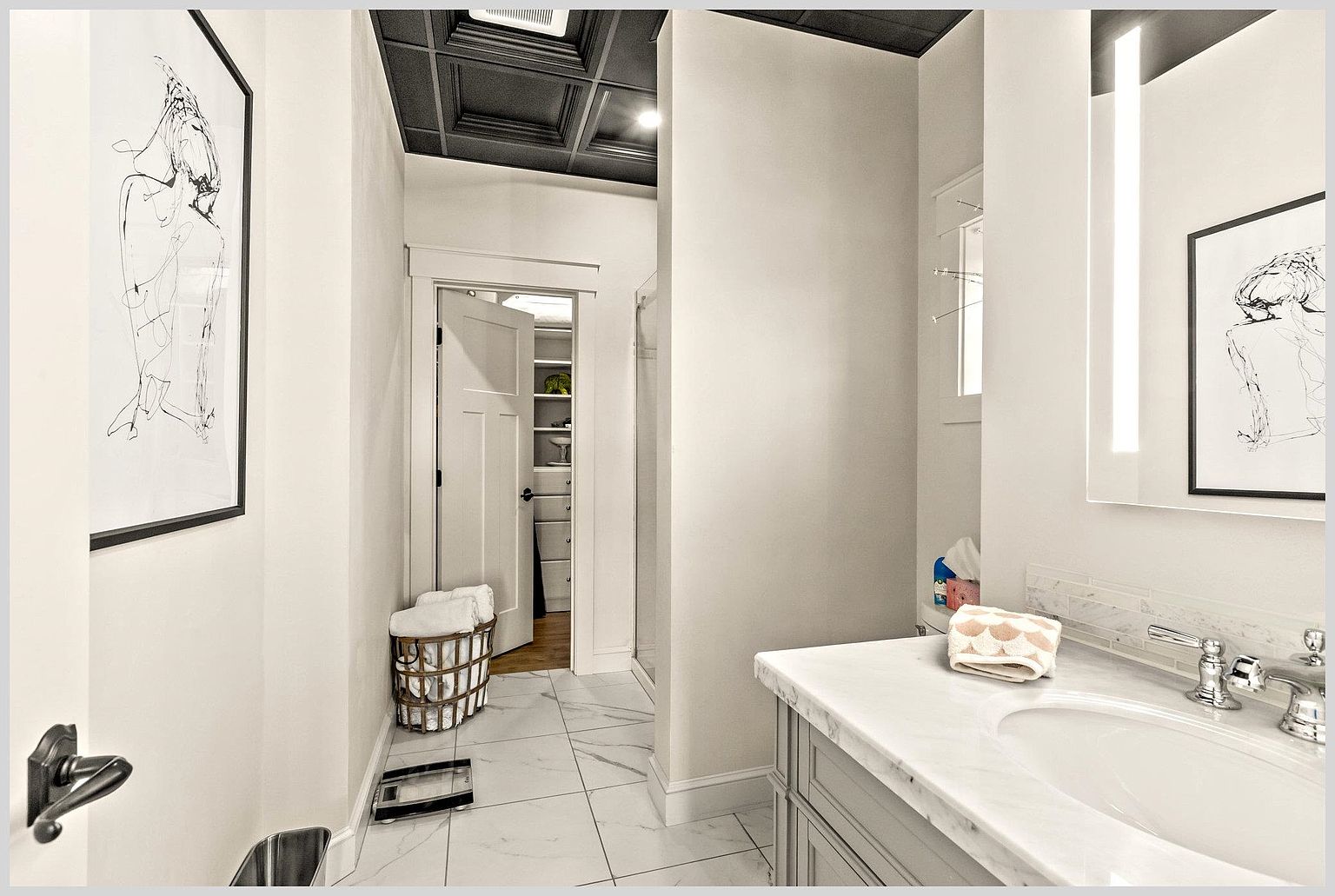 This bright bathroom features a vanity with a light-colored stone countertop and a modern sink fixture. The space includes a walk-in shower area and a doorway leading to a walk-in closet, all accented by a dark, coffered ceiling. The room is finished with large-format light-colored floor tiles and minimalist wall art, creating a clean and sophisticated aesthetic.