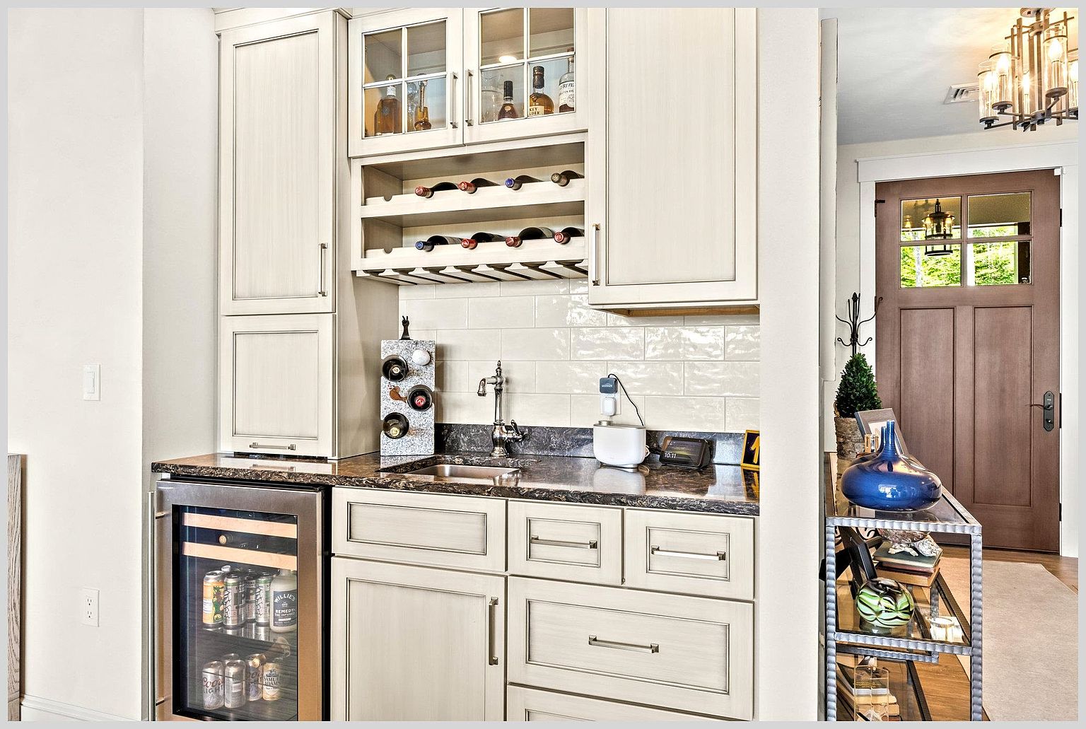 This image showcases a well-appointed wet bar area featuring light-toned cabinetry, a dark stone countertop, and a built-in beverage refrigerator. The space includes a small sink, a dedicated wine rack, and glass-front upper cabinets, all set against a glossy subway tile backsplash. To the right, a metal console table with a blue decorative vase sits near a wooden front door, creating a welcoming transition area.