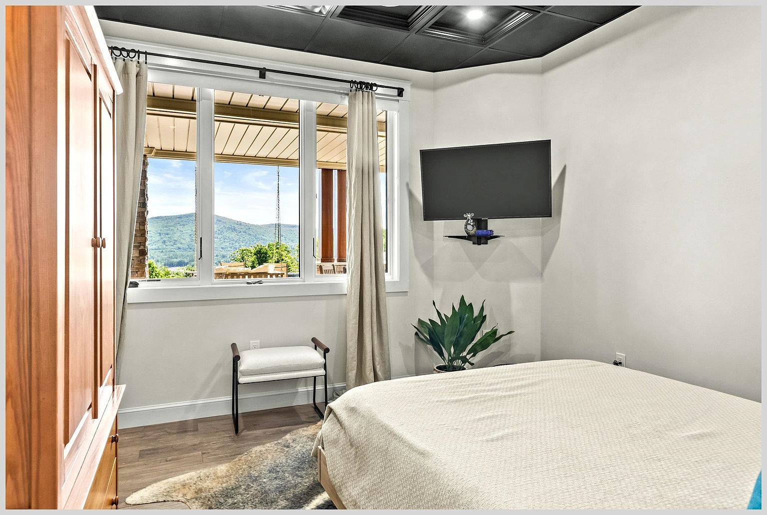 This bedroom features a neutral color palette with light walls and a dark, coffered ceiling that adds architectural interest. A large window provides a scenic view of the mountains, while a wall-mounted television and a small bench offer functional convenience. The room is accented by a patterned area rug and a potted plant, creating a cozy and inviting atmosphere.