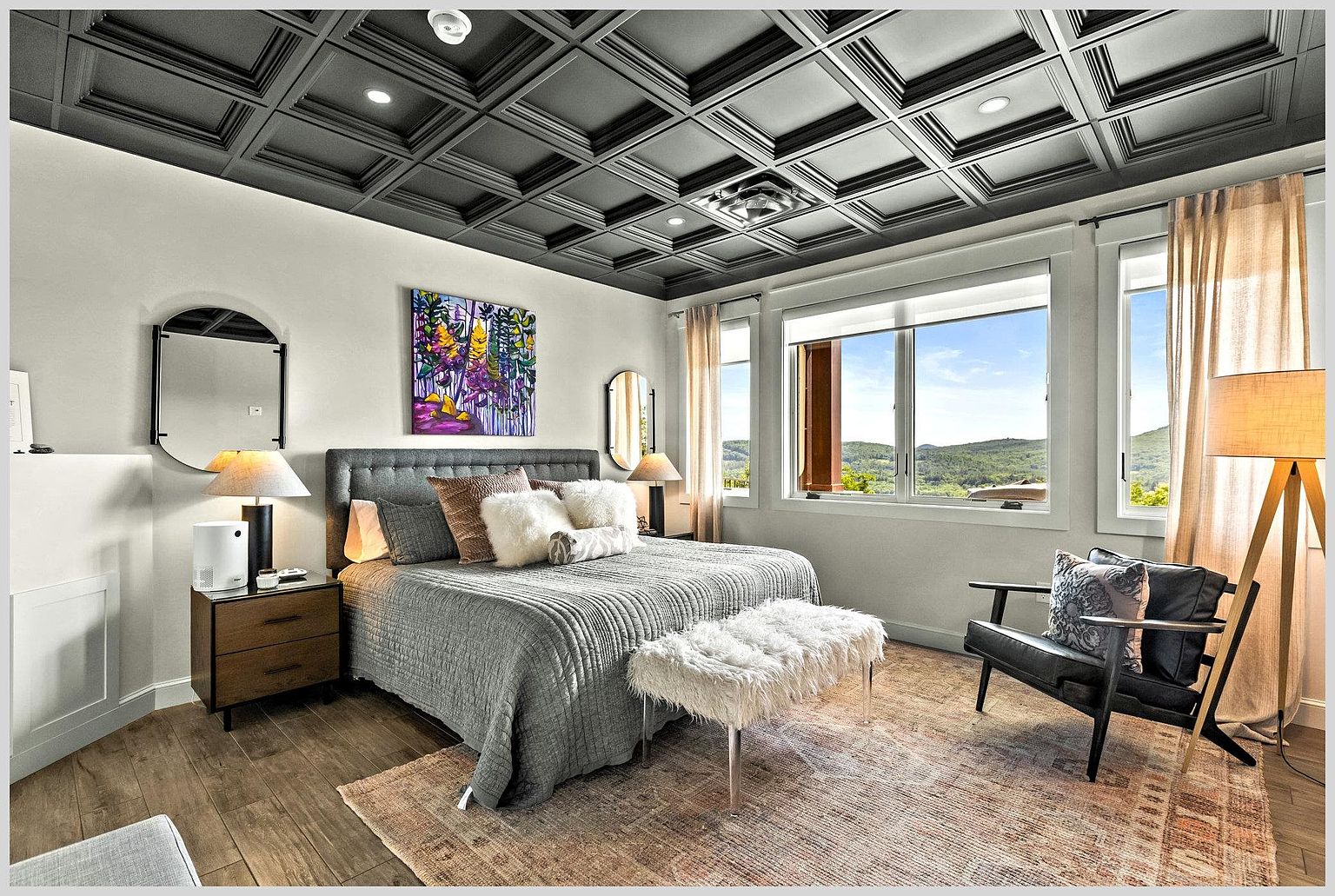 This primary bedroom features a striking coffered ceiling painted in a dark tone, contrasting with the light-colored walls and wood-look flooring. A plush, tufted-headboard bed serves as the focal point, complemented by a textured rug, a faux-fur bench, and a modern armchair near the large windows that frame a scenic mountain view. The room is accented by symmetrical mirrors, bedside lamps, and a vibrant piece of abstract wall art, creating a sophisticated and serene atmosphere.