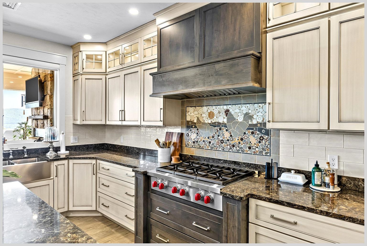 This well-appointed kitchen features a blend of light-toned cabinetry and a dark, stained wood range hood and base cabinet, creating a sophisticated contrast. A prominent mosaic tile backsplash depicting chickens serves as a focal point above the professional-grade gas range, while dark stone countertops provide a rich, textured surface throughout. The space is illuminated by recessed lighting and offers a view through a window toward an outdoor stone feature, emphasizing a warm and inviting atmosphere.