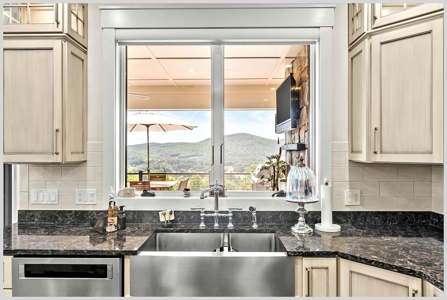 This kitchen features a large stainless steel farmhouse sink set into a dark, patterned stone countertop, flanked by light-toned cabinetry. A central window provides a clear, scenic view of a mountain landscape and an outdoor patio area with an umbrella. The space is finished with a neutral subway tile backsplash and includes a decorative glass cloche on the counter, creating a bright and inviting atmosphere.