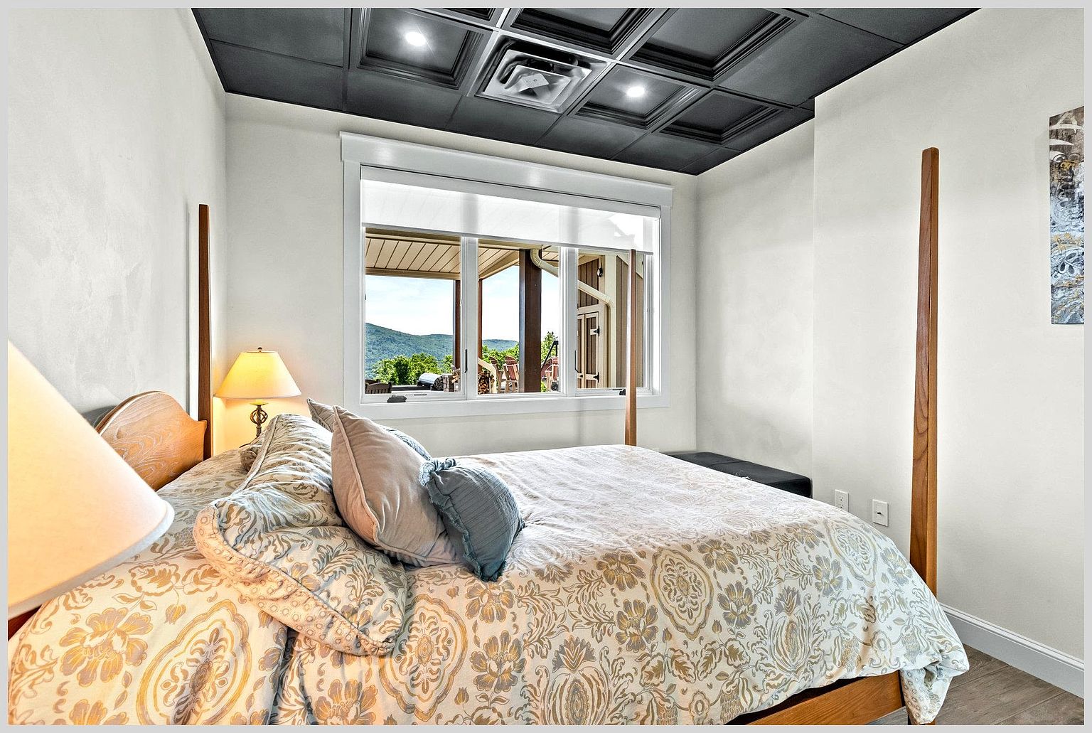 This bedroom features a four-poster bed with a patterned beige and gold comforter, positioned beneath a distinctive black coffered ceiling. A large window provides a view of the outdoor deck and surrounding mountain landscape, filling the room with natural light. The space is accented by a warm bedside lamp and minimalist wall decor, creating a cozy and inviting atmosphere.