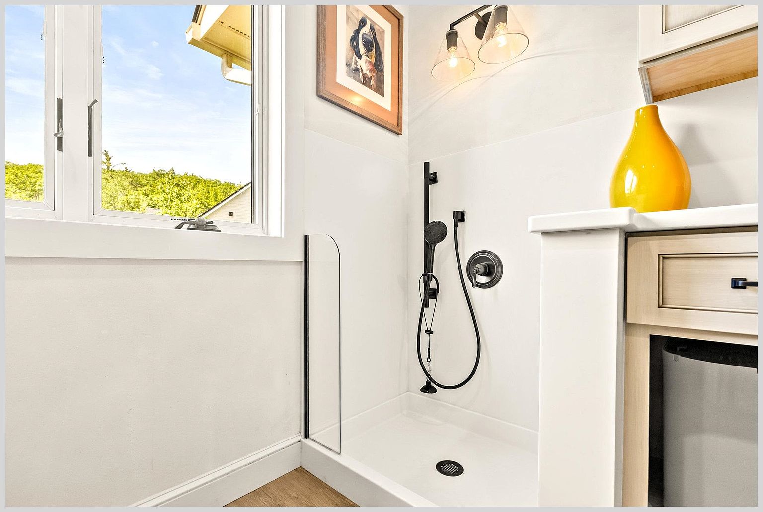 This bright bathroom features a walk-in shower with a glass panel, matte black fixtures, and a handheld showerhead. To the right, a vanity with light-toned cabinetry is topped with a yellow decorative vase, while a framed piece of art hangs on the wall above the shower. A large window provides natural light, creating an airy and clean aesthetic.