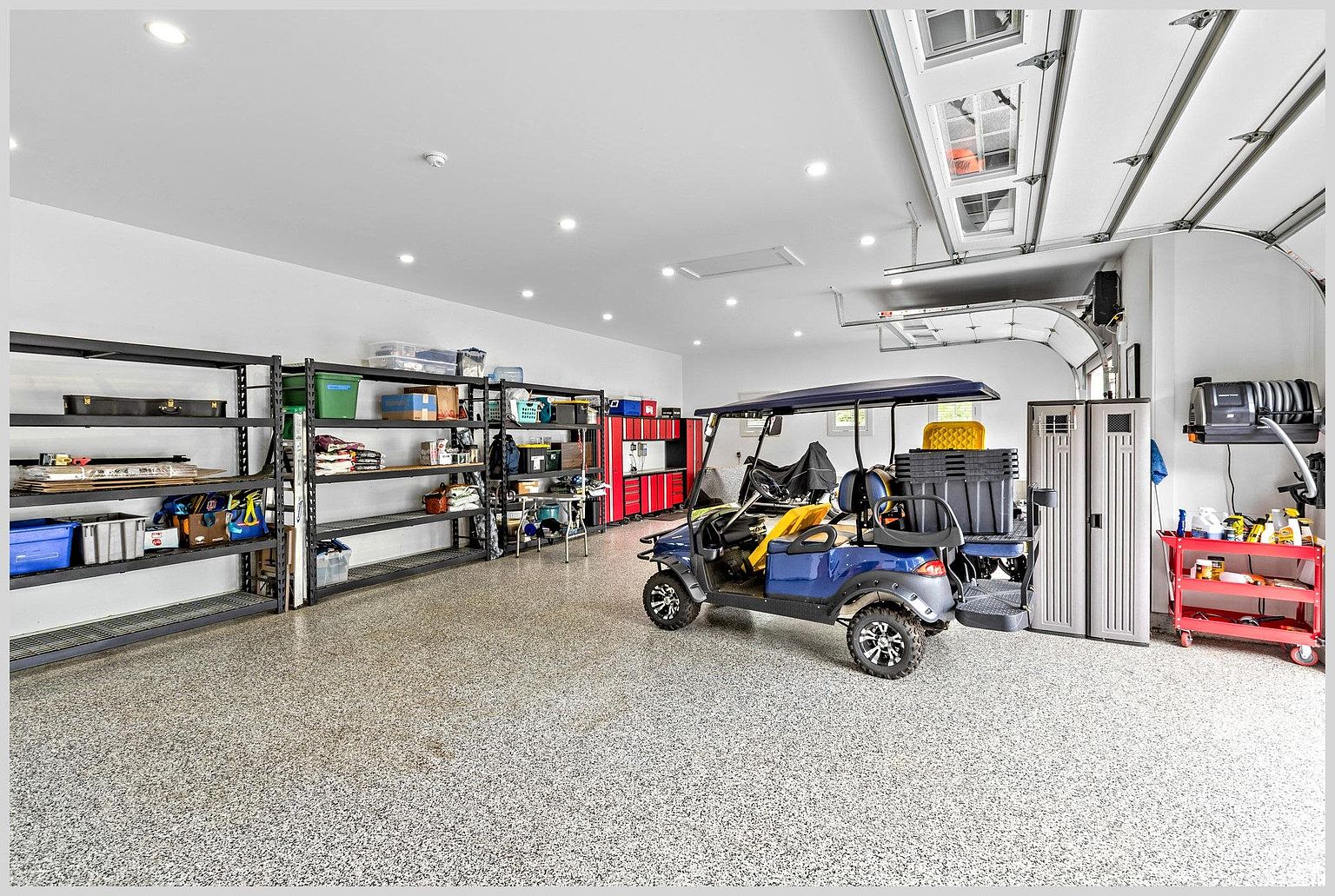 This spacious, well-organized garage features a durable speckled epoxy floor and bright recessed lighting. The area is equipped with extensive black metal shelving units for storage, a red rolling tool cart, and a blue golf cart parked in the center. The perspective is a wide-angle shot from the garage entrance, showcasing the clean, functional layout and ample storage capacity.