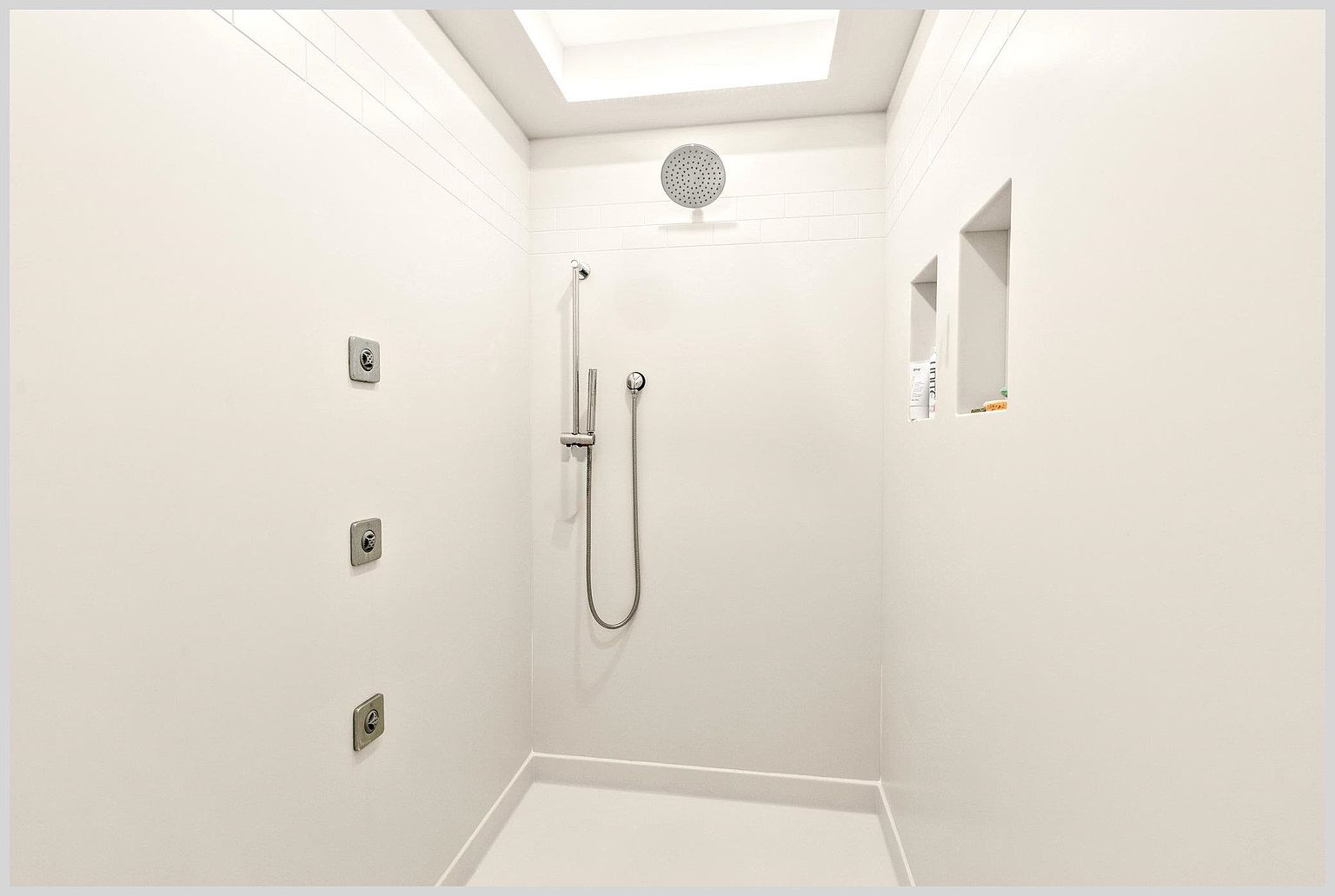 This minimalist walk-in shower features a clean, all-white aesthetic with a large rainfall showerhead mounted on the ceiling and a separate handheld sprayer. The space includes three integrated wall-mounted body sprayers and two recessed wall niches for storage, all set against smooth, seamless walls. A skylight above provides natural illumination, creating a bright and airy atmosphere.