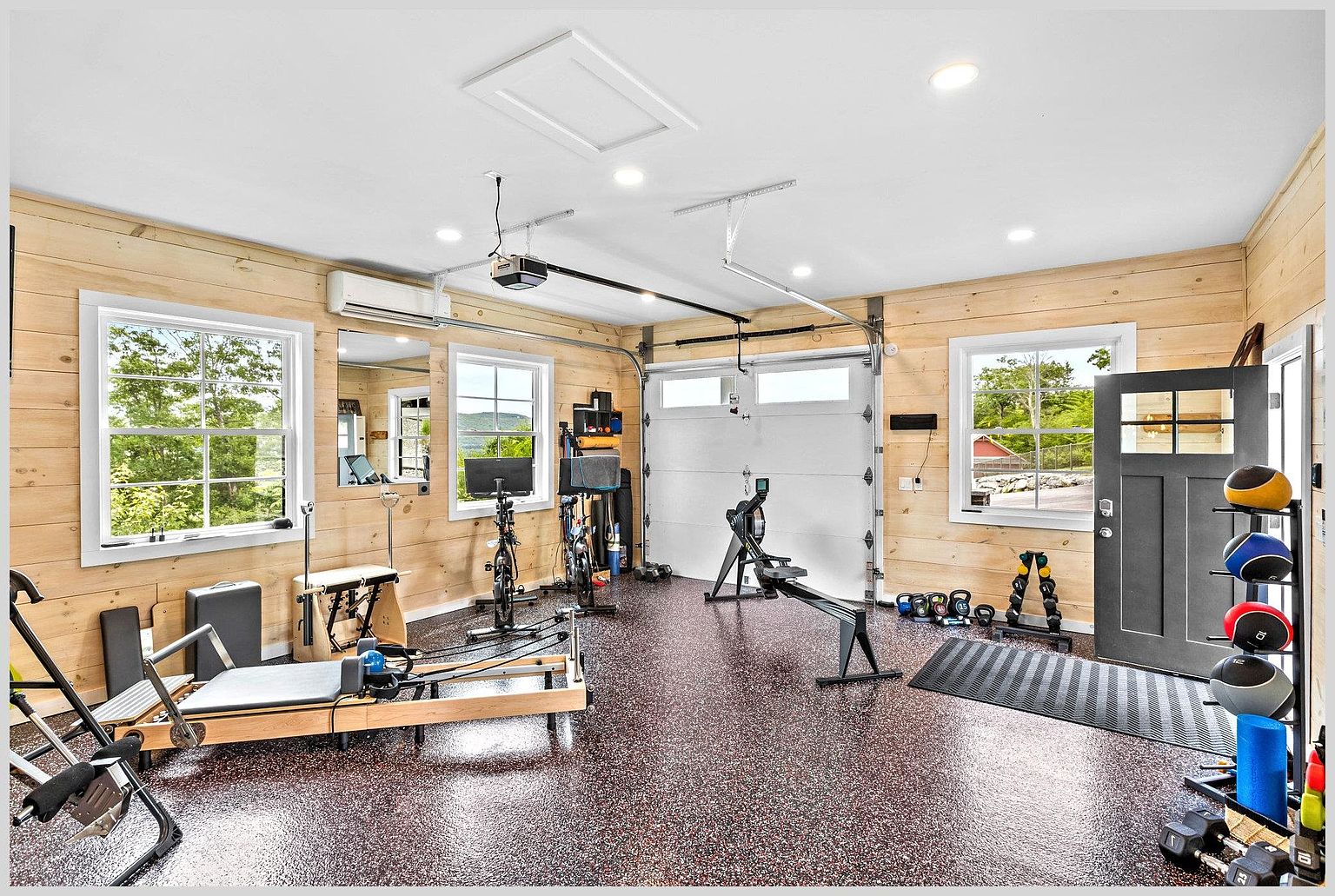 This well-equipped home gym features light wood-paneled walls and durable, speckled flooring, creating a bright and functional workout space. The room is outfitted with a variety of fitness equipment, including a Pilates reformer, a rowing machine, stationary bikes, and a rack of medicine balls. Large windows provide natural light and scenic views, while recessed lighting ensures the space remains well-lit for exercise.