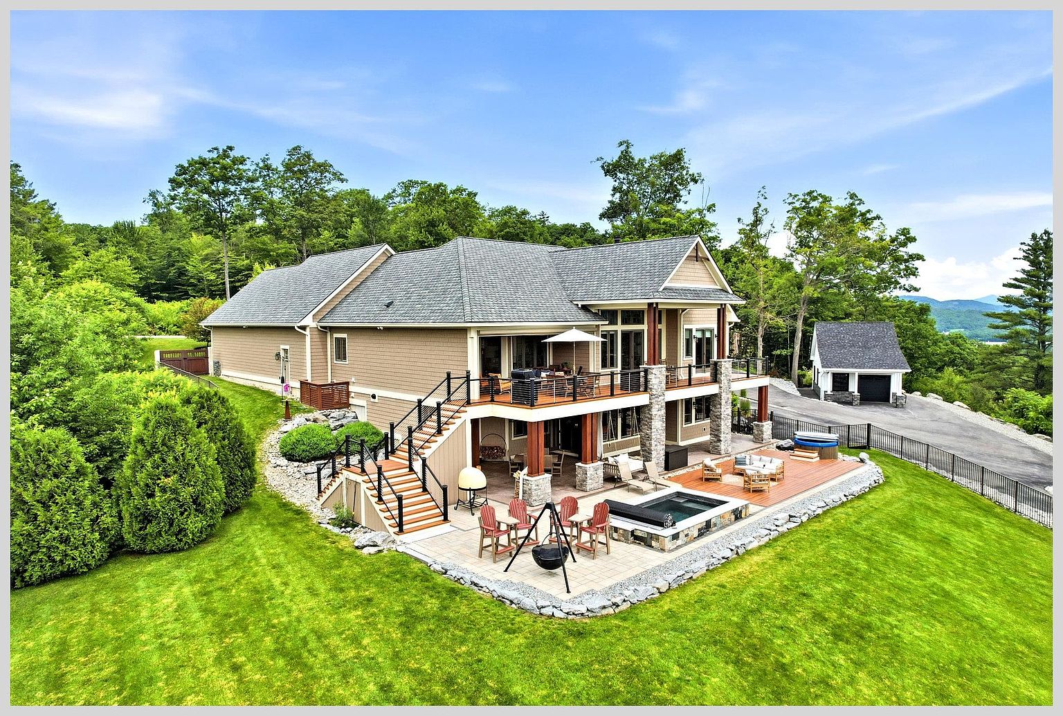 This elevated rear view captures a multi-level home featuring a spacious upper deck and a lower stone-paved patio area. The outdoor space is well-appointed with a built-in spa, a fire pit, and comfortable seating, all overlooking a lush green lawn and surrounding forest. The home's exterior combines neutral siding with stone accents, creating a warm and inviting aesthetic perfect for outdoor entertaining.