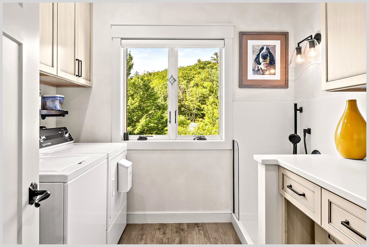 This bright and functional laundry room features white cabinetry, a white washer and dryer set, and a light-toned countertop with a modern black faucet. A central window provides a clear view of the lush green trees outside, while a framed dog portrait adds a personalized touch to the wall. The space is finished with wood-look flooring and clean, neutral walls, creating a tidy and inviting atmosphere.