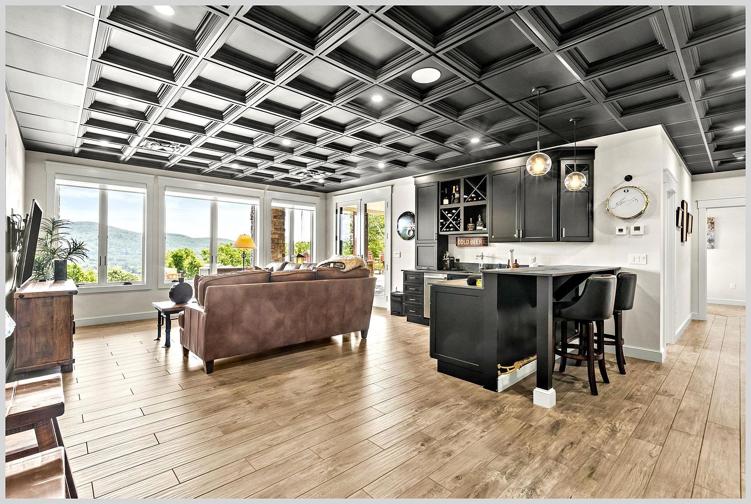 This open-concept living area features a striking coffered ceiling painted in a dark tone, contrasting with the light wood-look flooring. A comfortable brown sofa faces a television, while a built-in wet bar with dark cabinetry and pendant lighting sits to the side. Large windows provide an expansive view of the surrounding mountains, creating a bright and inviting atmosphere.