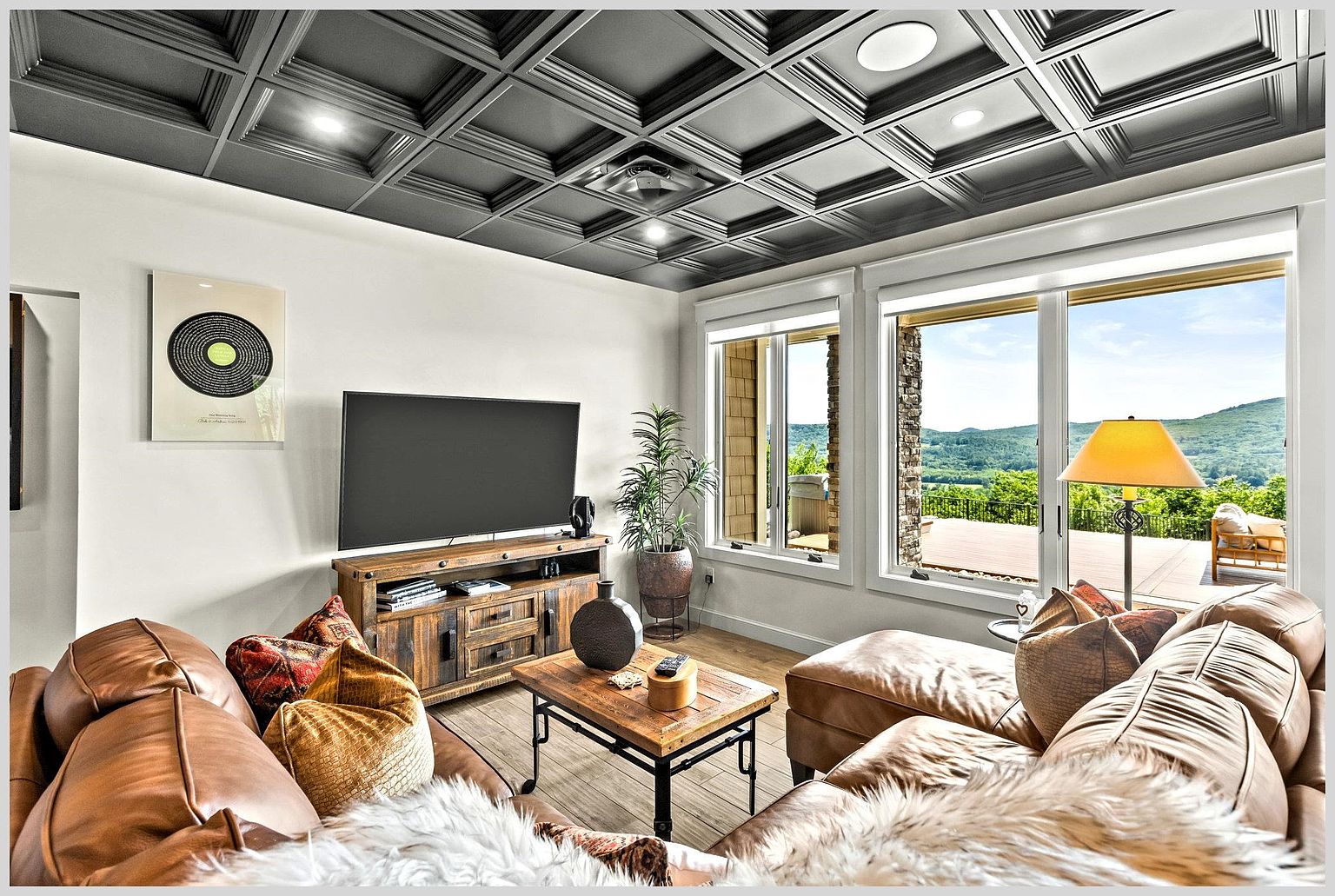 This inviting living room features a rich, dark coffered ceiling that adds architectural depth to the space. A large leather sectional sofa and a rustic wood media console create a cozy atmosphere, complemented by a central coffee table and a decorative record-themed wall art piece. Large windows provide an expansive view of the surrounding mountains and a wooden deck, bathing the room in natural light.