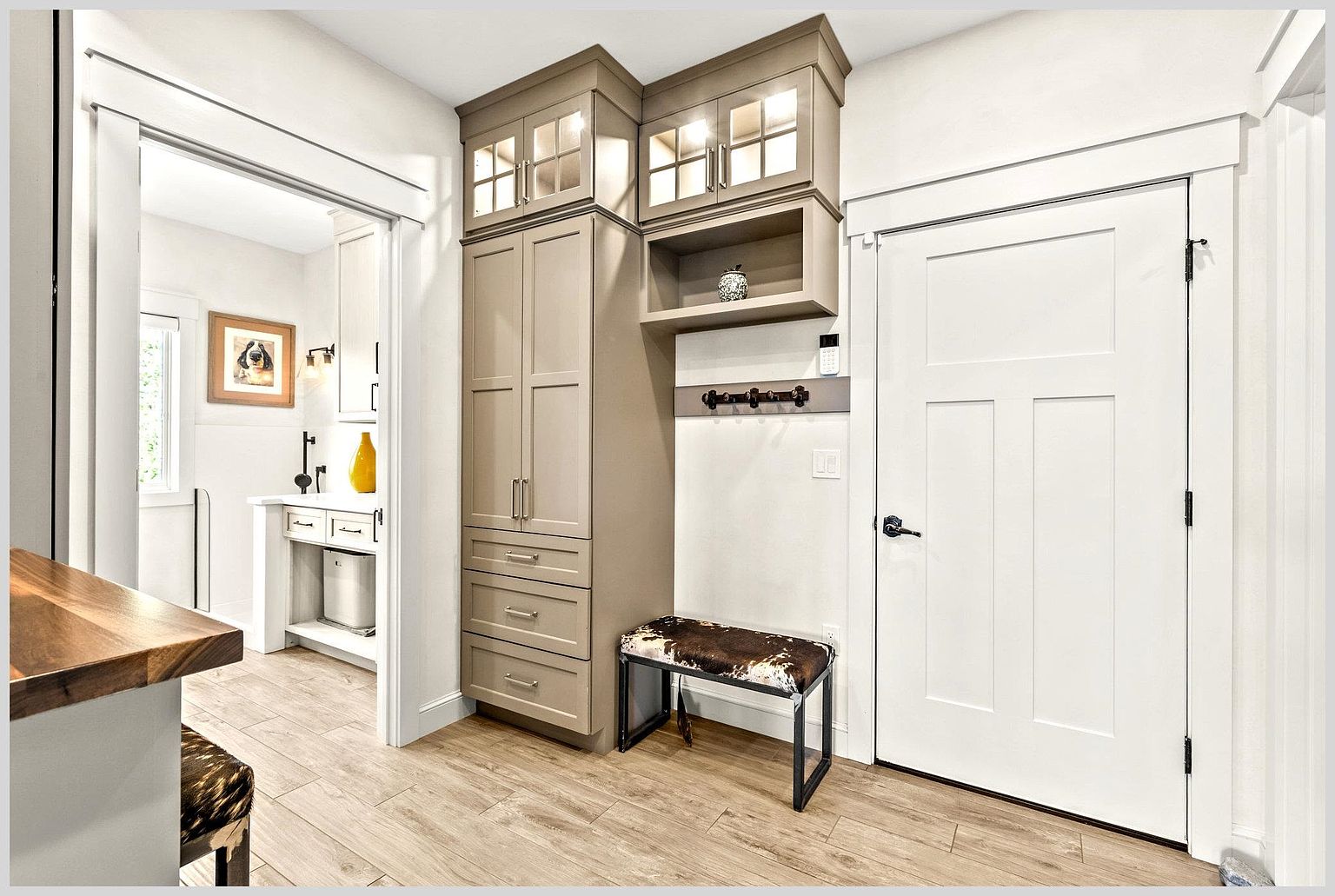 This mudroom area features custom taupe cabinetry with glass-front upper doors and a built-in bench with a cowhide-patterned cushion. To the right, a white door leads to another room, while an open doorway on the left provides a glimpse into a bright bathroom with a vanity and framed artwork. The space is finished with light wood-look flooring and clean white trim, creating a functional and organized transition space.