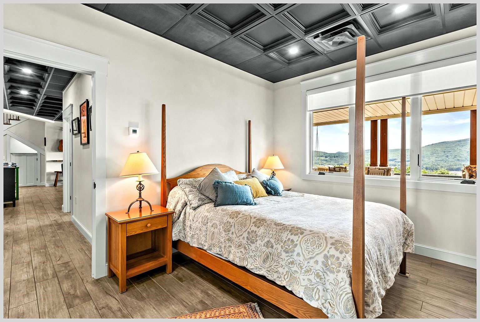 This cozy bedroom features a four-poster wooden bed frame centered in the room, dressed in a patterned comforter with blue and yellow accent pillows. The space is highlighted by a dark, coffered ceiling and light wood-look flooring, with large windows providing a scenic view of the surrounding mountains. A wooden nightstand with a lamp sits to the left, while a doorway leads into a bright, adjacent hallway.