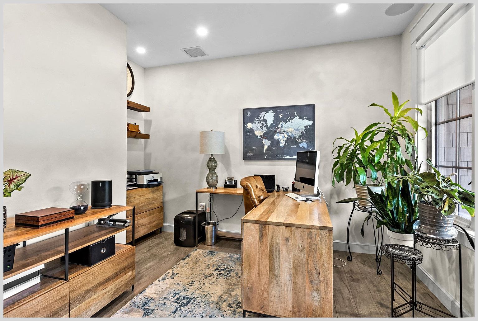 This well-appointed home office features a large wooden desk as the centerpiece, complemented by a leather chair and a patterned area rug. The room includes a dedicated workspace with a computer, a printer station on a wooden cabinet, and open shelving for decor. Large windows allow natural light to brighten the space, which is accented by several potted plants and a world map wall art.