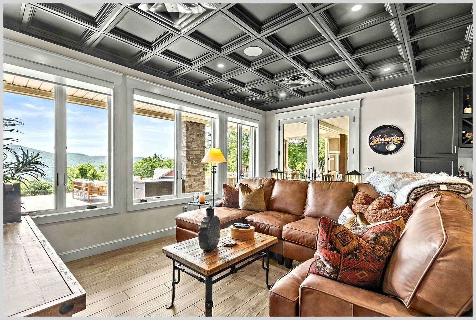 This inviting living room features a rich brown leather sectional sofa adorned with patterned throw pillows and a soft fur throw. The space is defined by a striking dark coffered ceiling and light wood-look flooring, with large windows providing expansive views of the surrounding mountains. A rustic wood and metal coffee table sits at the center, while a circular sign adds a personalized touch to the wall near the glass doors leading to the outdoor deck.