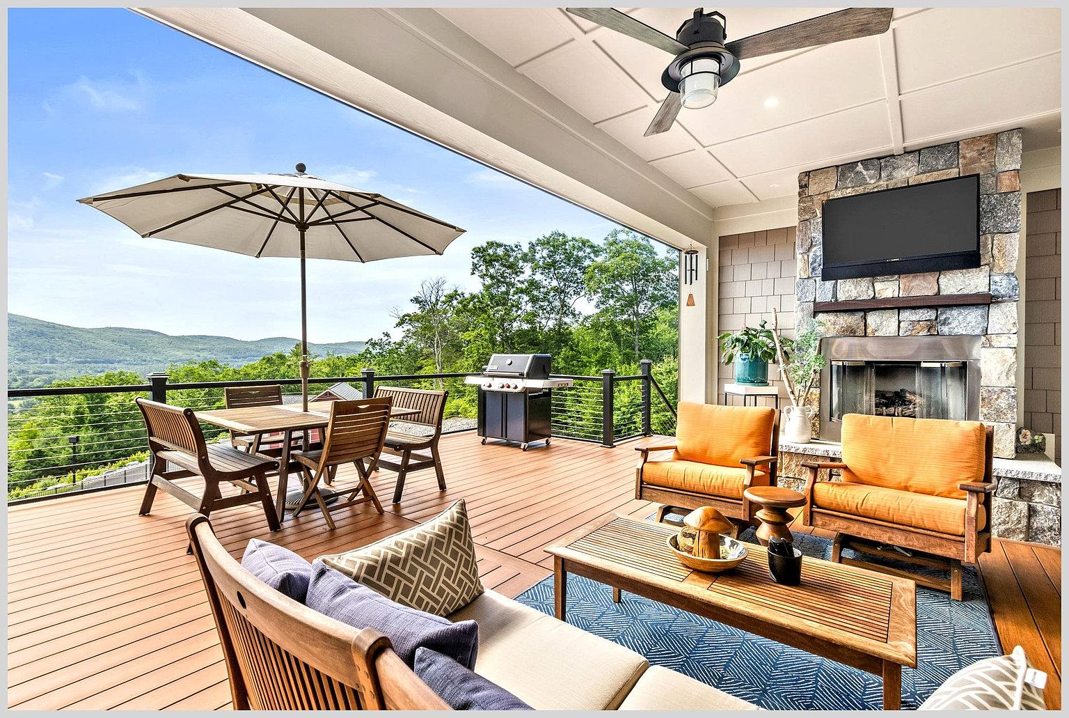 This expansive covered deck offers a seamless transition between indoor and outdoor living, featuring a stone-clad chimney breast with an integrated television and fireplace. The space is furnished with comfortable lounge seating and a dining area, all overlooking a scenic mountain landscape. The deck is constructed with warm-toned decking material and includes a ceiling fan for comfort, creating an inviting atmosphere for relaxation or entertaining.