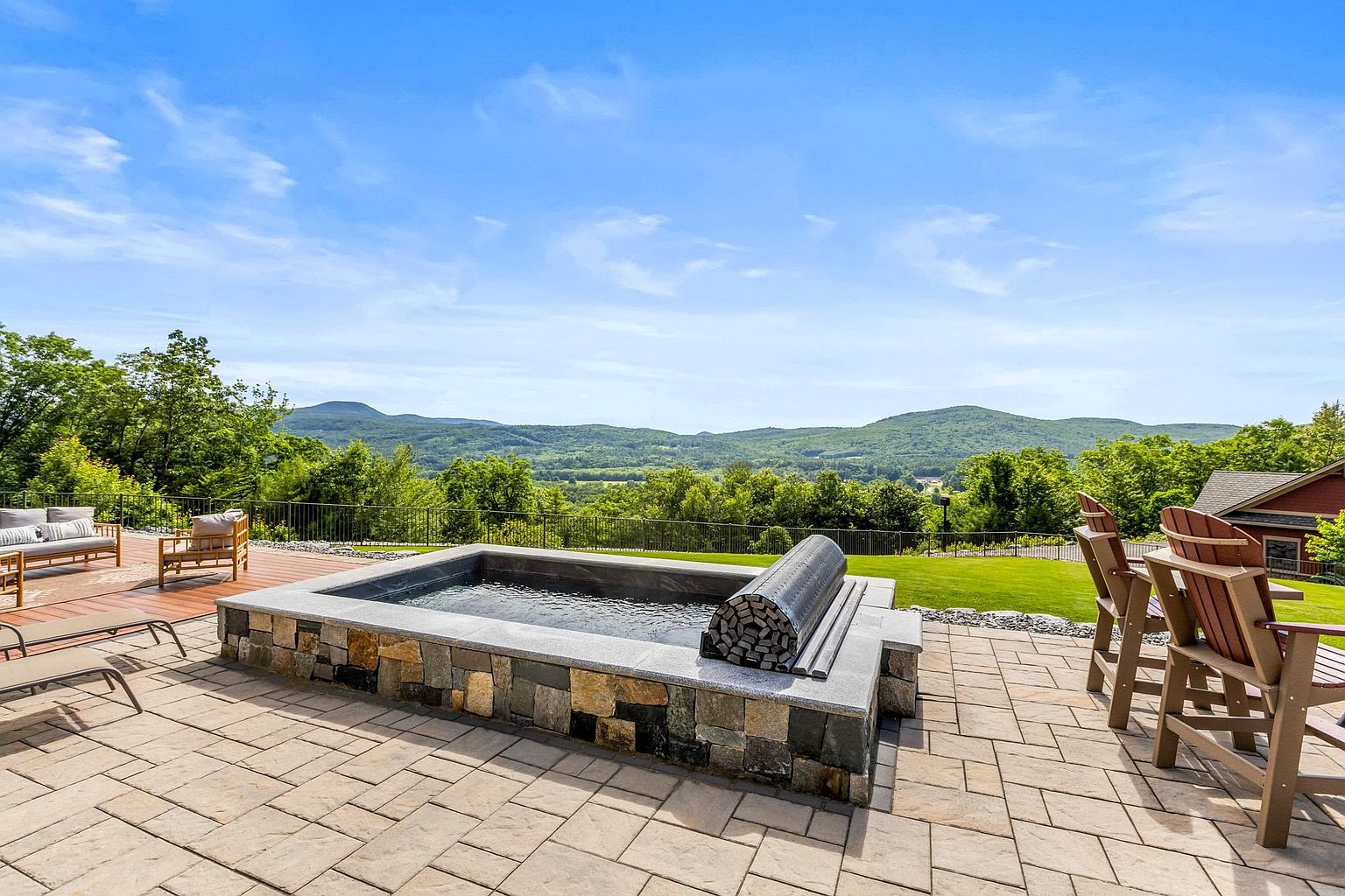 This expansive stone-paved patio features a custom-built, stone-clad spa as its centerpiece, complete with a retractable cover. The outdoor living space is furnished with comfortable lounge seating and Adirondack chairs, all oriented to take advantage of the sweeping, panoramic mountain views. The setting is serene and upscale, highlighting a seamless transition between luxury outdoor amenities and the natural landscape.