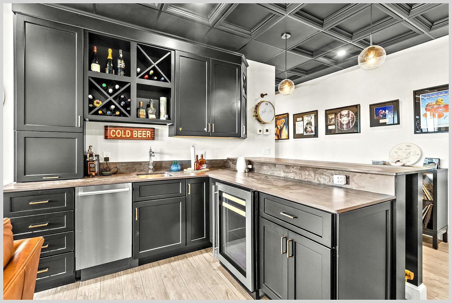 This sophisticated home bar area features dark, matte-finish cabinetry paired with a neutral-toned countertop and a coffered ceiling. The space is equipped with a stainless steel dishwasher, a built-in wine refrigerator, and a dedicated wine rack, creating an ideal setup for entertaining. The perspective captures the L-shaped layout, highlighting the sleek hardware and the decorative framed art on the adjacent wall.