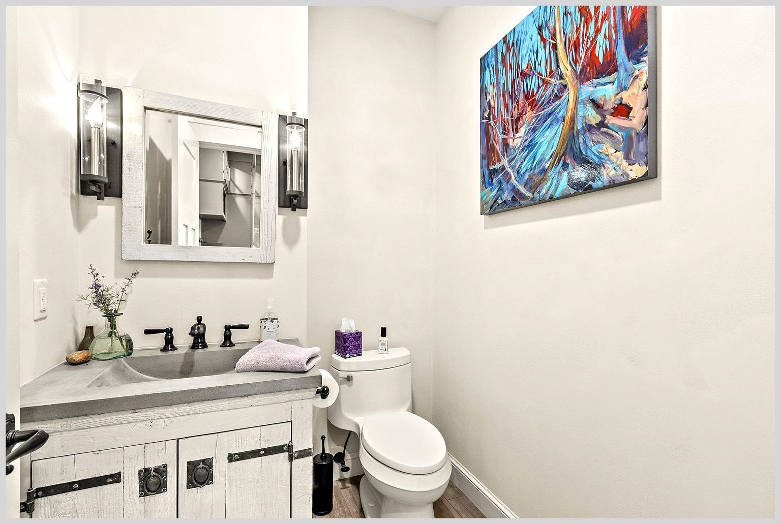 This compact powder room features a rustic-style vanity with a concrete-look countertop and a distressed white wood cabinet. A rectangular mirror with a weathered frame is flanked by two industrial-style wall sconces, while a vibrant, abstract painting adds a pop of color to the neutral wall. The space is completed by a standard white toilet and a small decorative vase, creating a cozy and character-filled atmosphere.